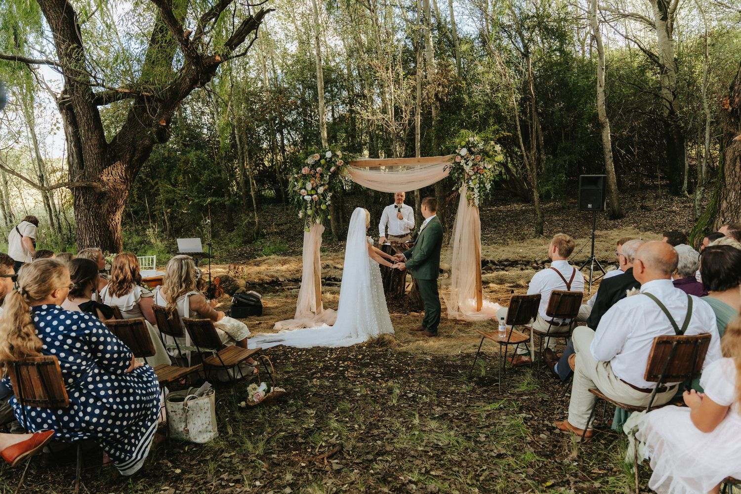Outdoor woodland wedding ceremony with couple at floral arch altar, guests seated on chairs among trees.