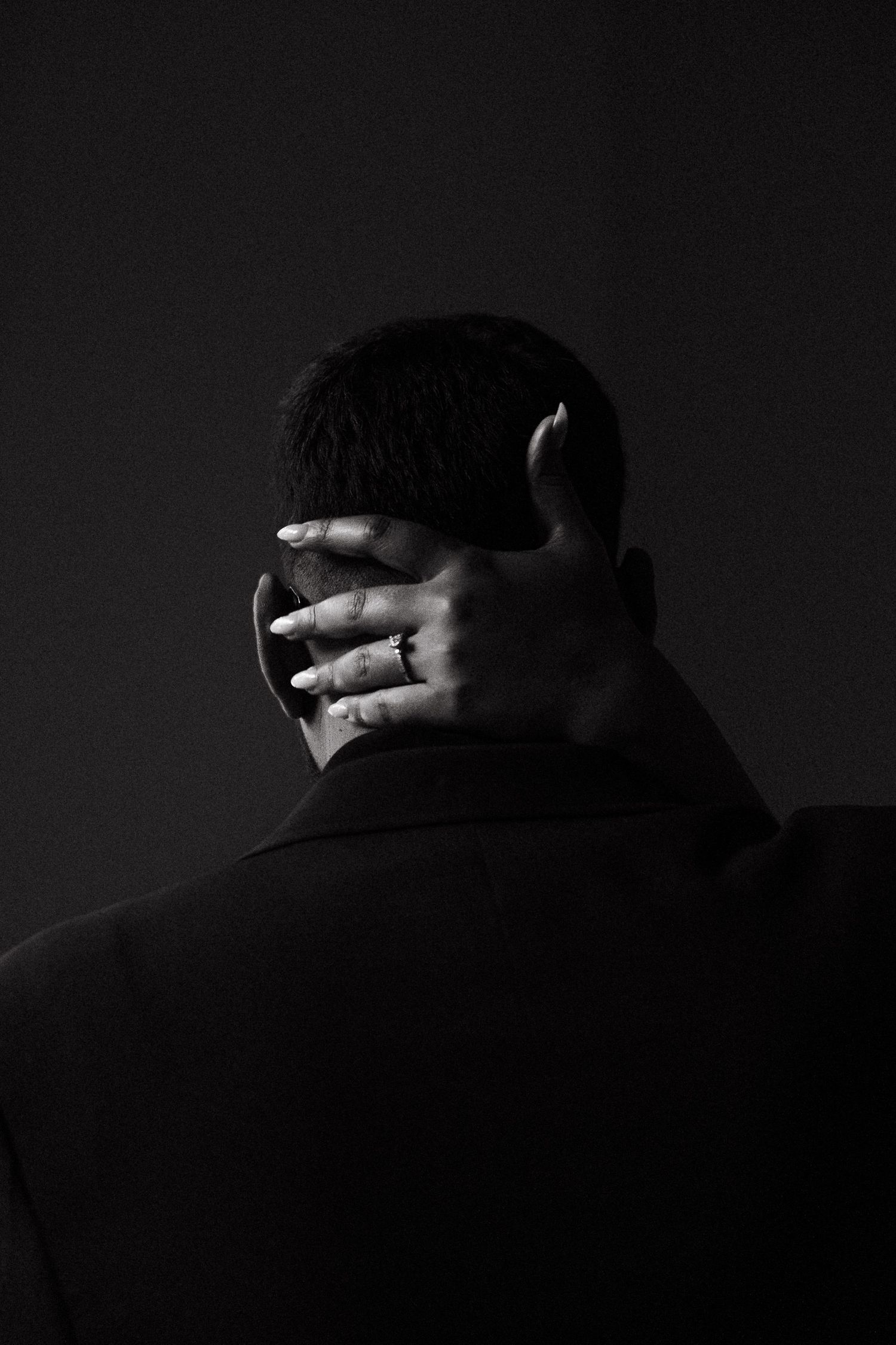 Black and white photo of a man seen from behind with a hand resting on his neck, wearing a ring.