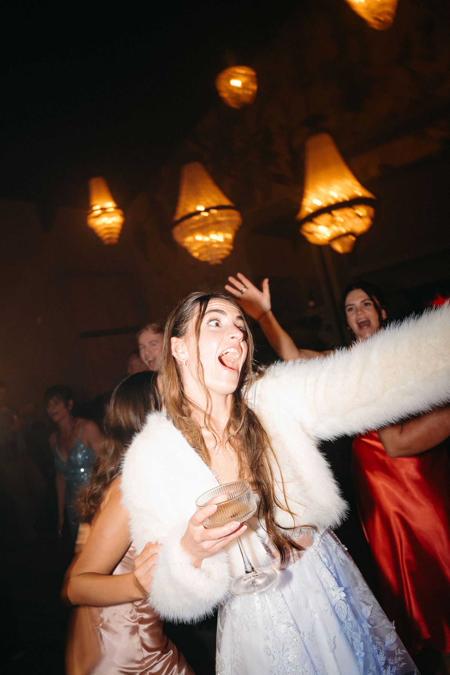 Bride in white dress and fur shawl dancing joyfully with champagne glass at elegant wedding reception in Plettenberg Bay.