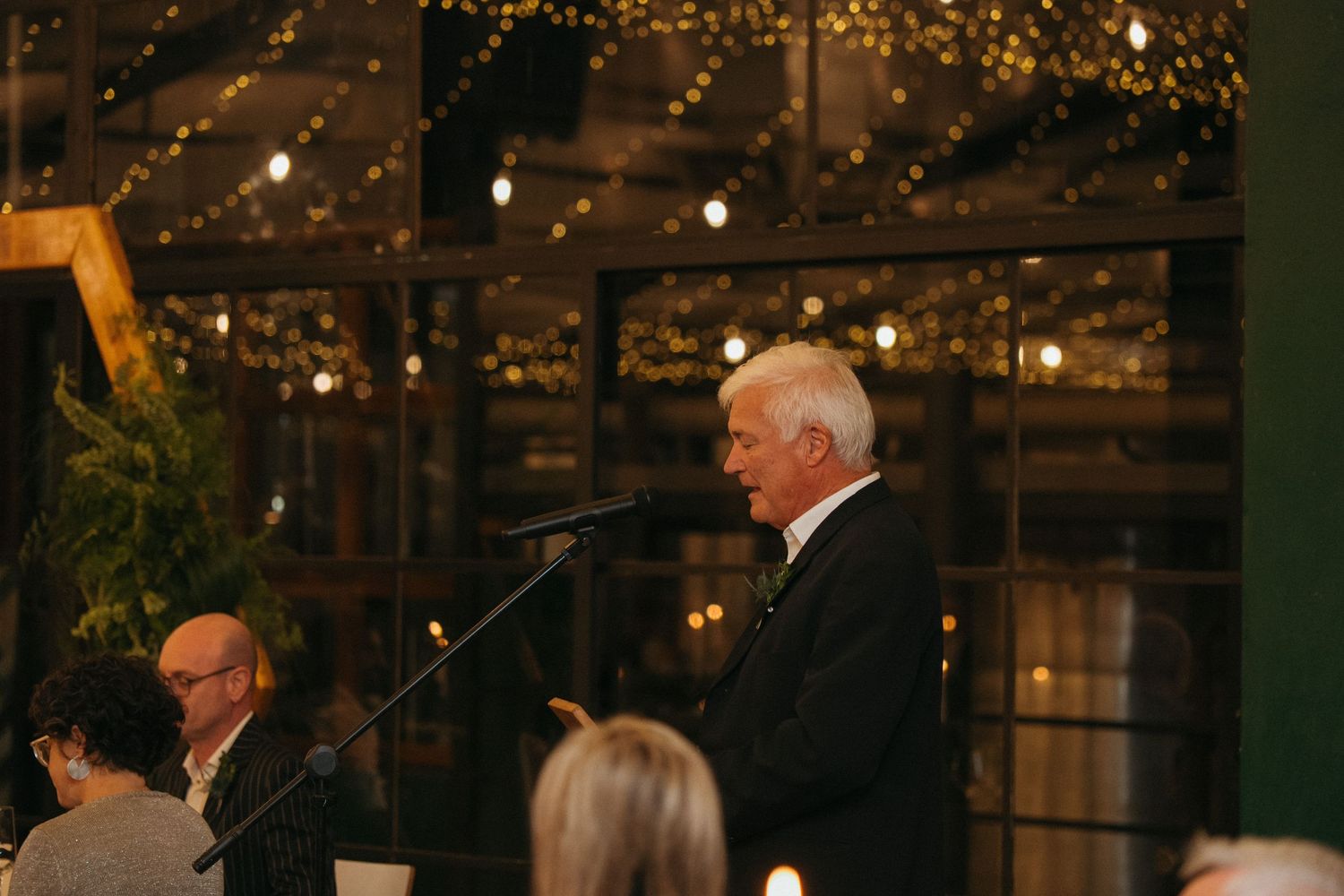 Elderly man in dark suit speaking at podium during evening event with warm string lights in background.