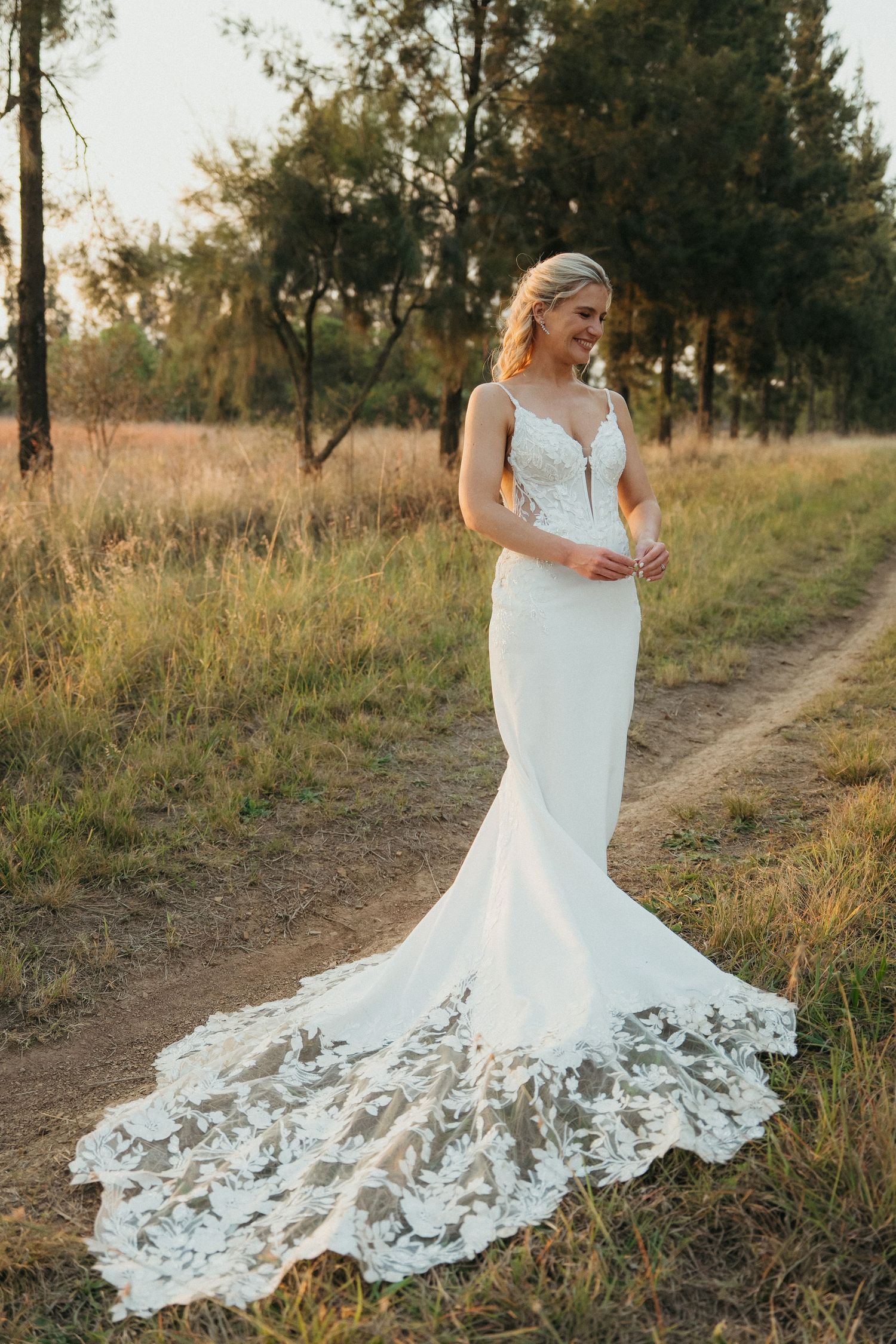 Bride in a fitted lace mermaid wedding gown with a long train stands on a dirt path surrounded by golden grass.