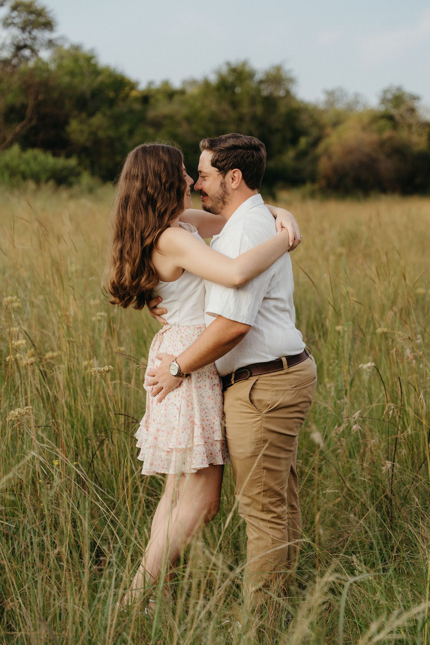 Couple embracing in a golden meadow, woman in floral dress and man in white shirt, sharing a romantic moment outdoors.