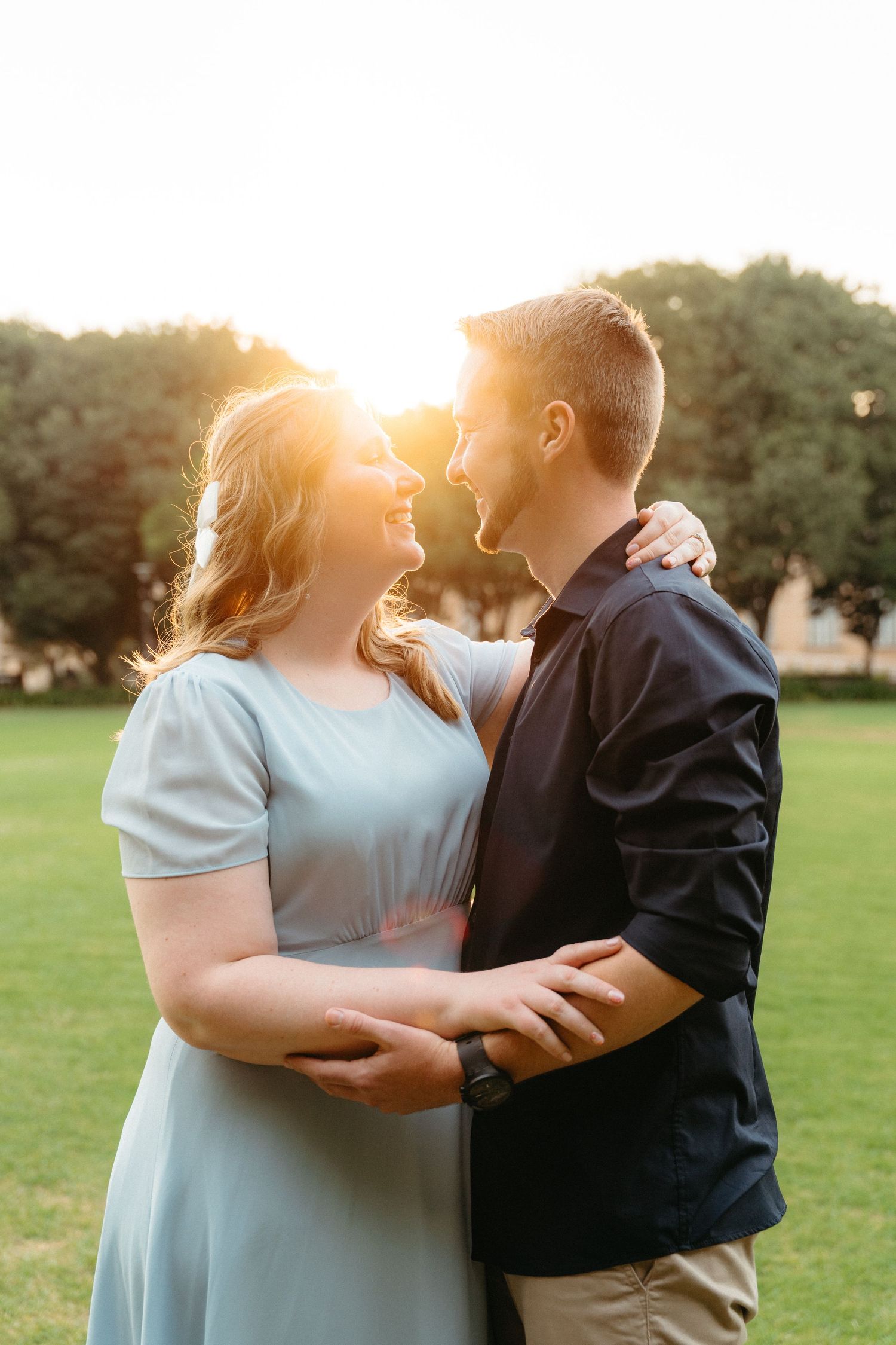 A romantic couple embraces in a grassy field during golden hour sunset with sun flare behind them.