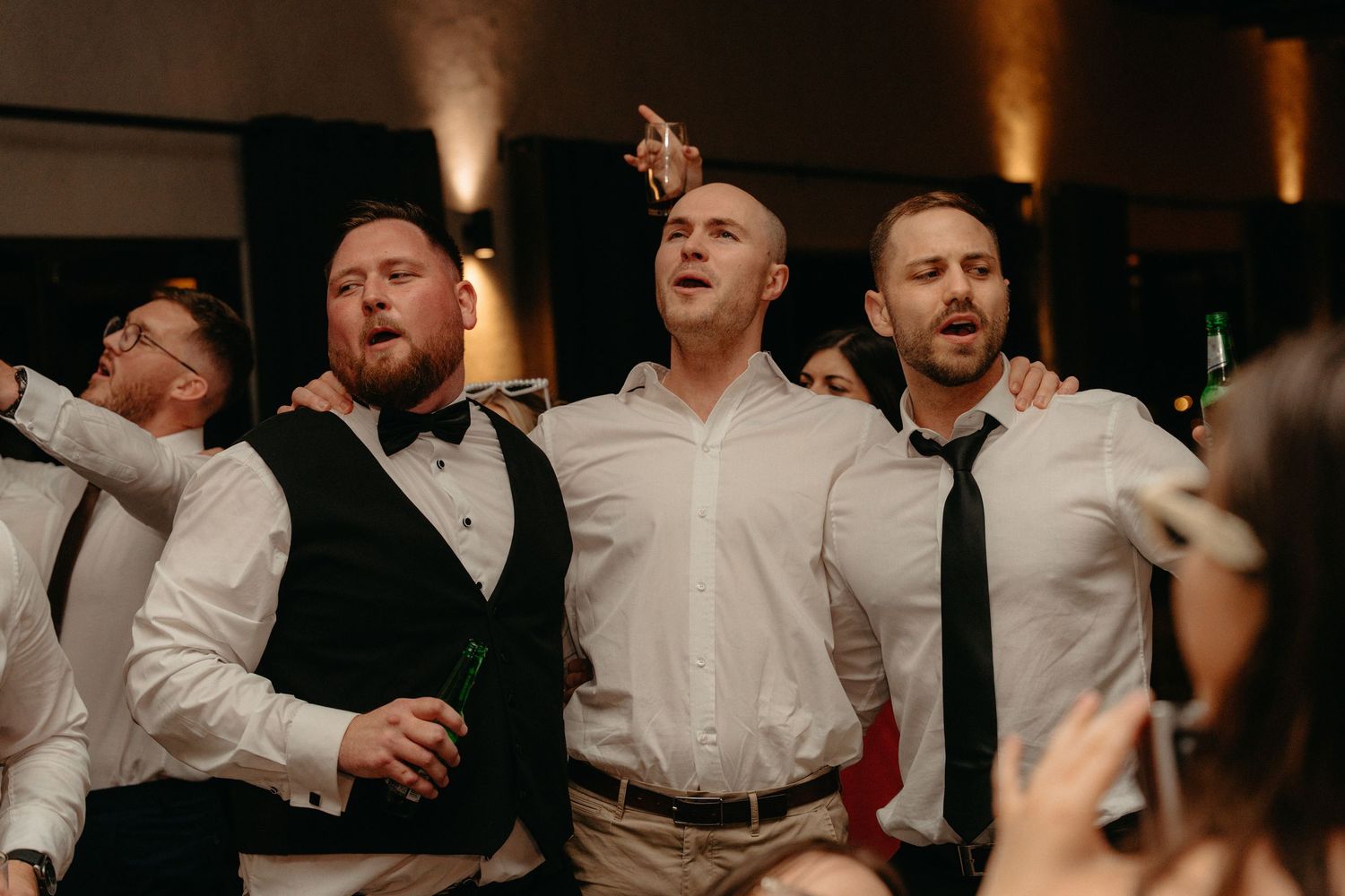 A group of groomsmen in white shirts and black ties dance enthusiastically at a wedding reception.