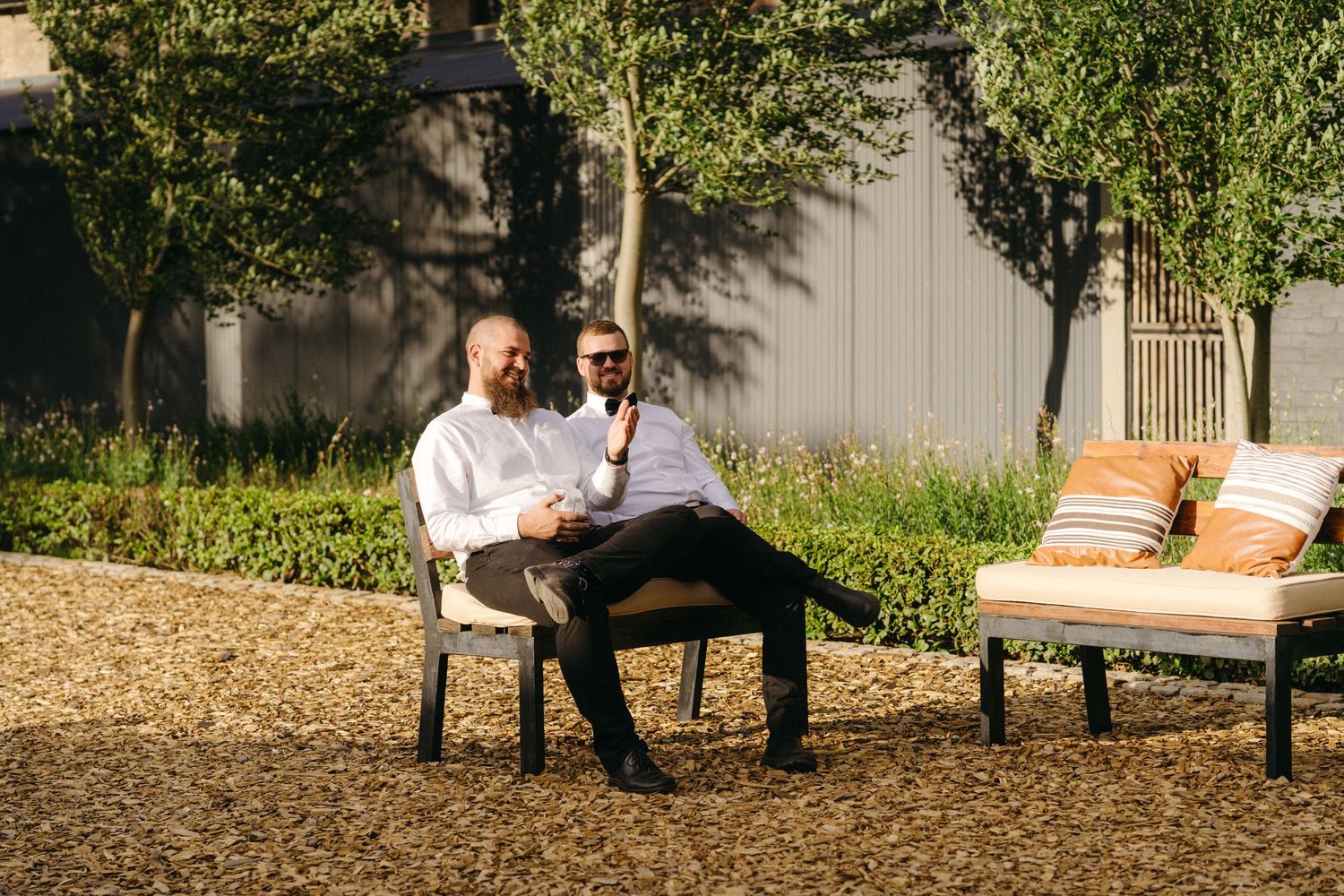 Two people sitting outdoors on patio furniture share a relaxed conversation in a garden setting with trees and cushions.