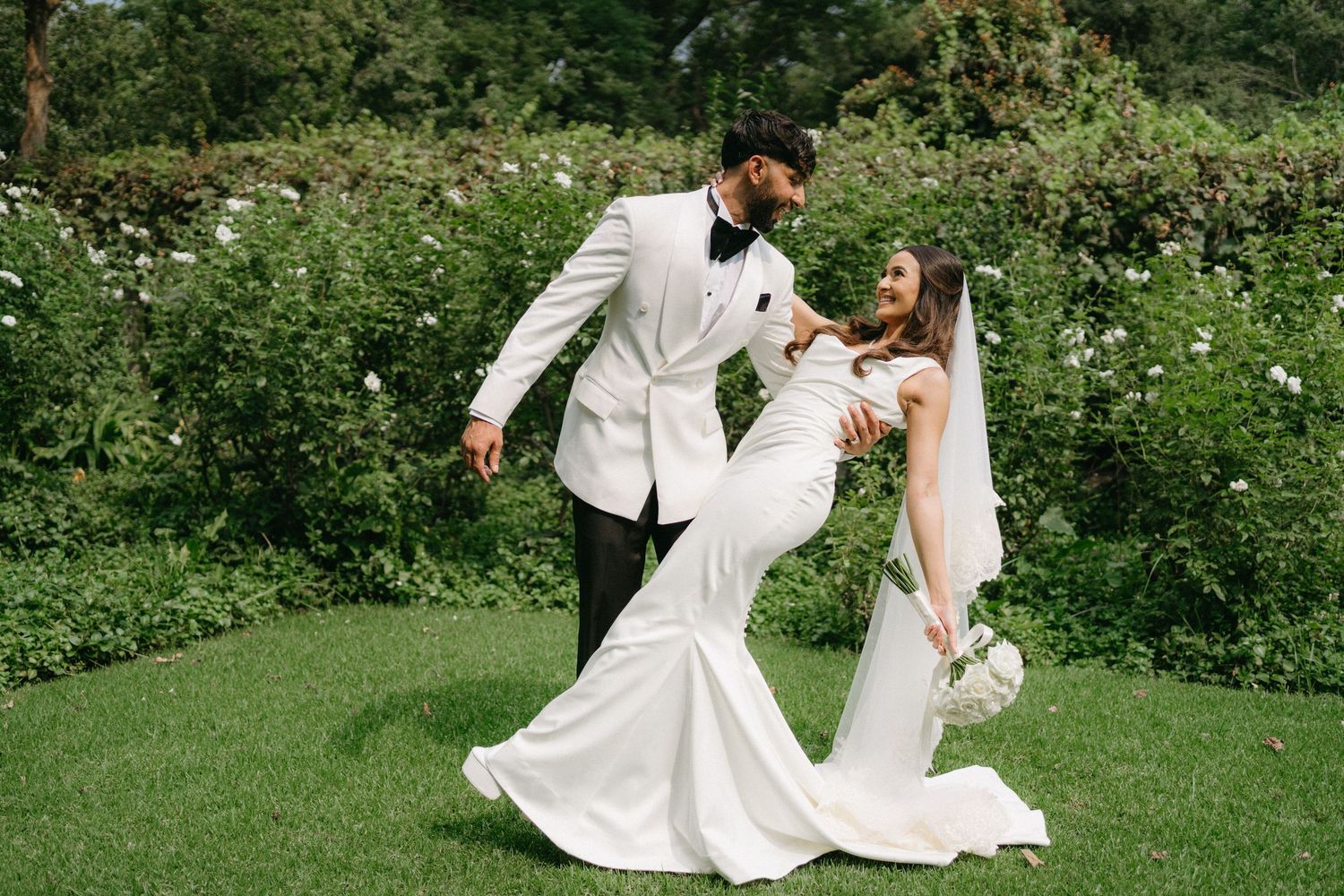 Couple in elegant white wedding attire pose romantically in a lush garden setting with wildflowers.