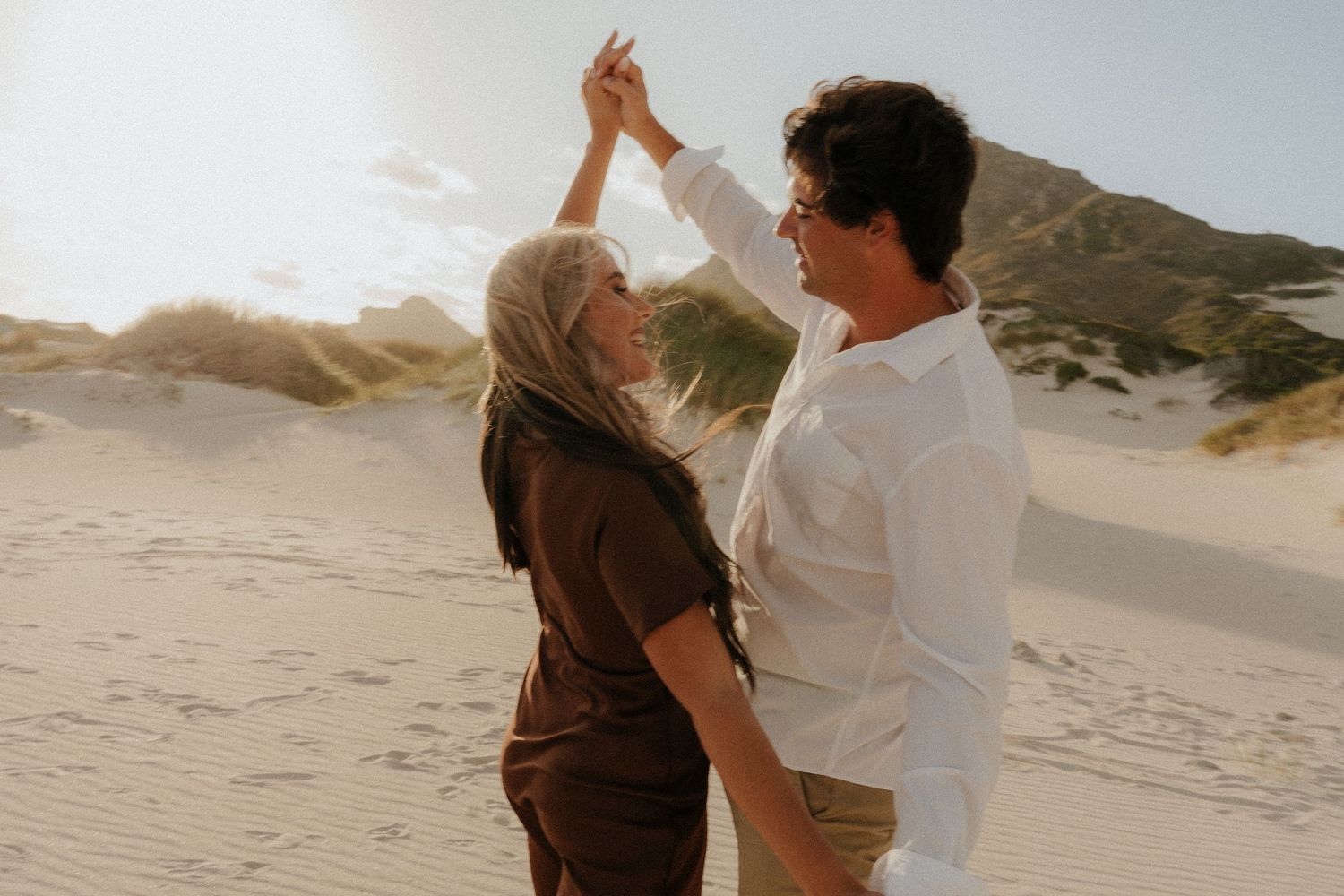 Couple dancing joyfully on sandy dunes, woman in brown dress, man in white shirt, sunny coastal landscape.
