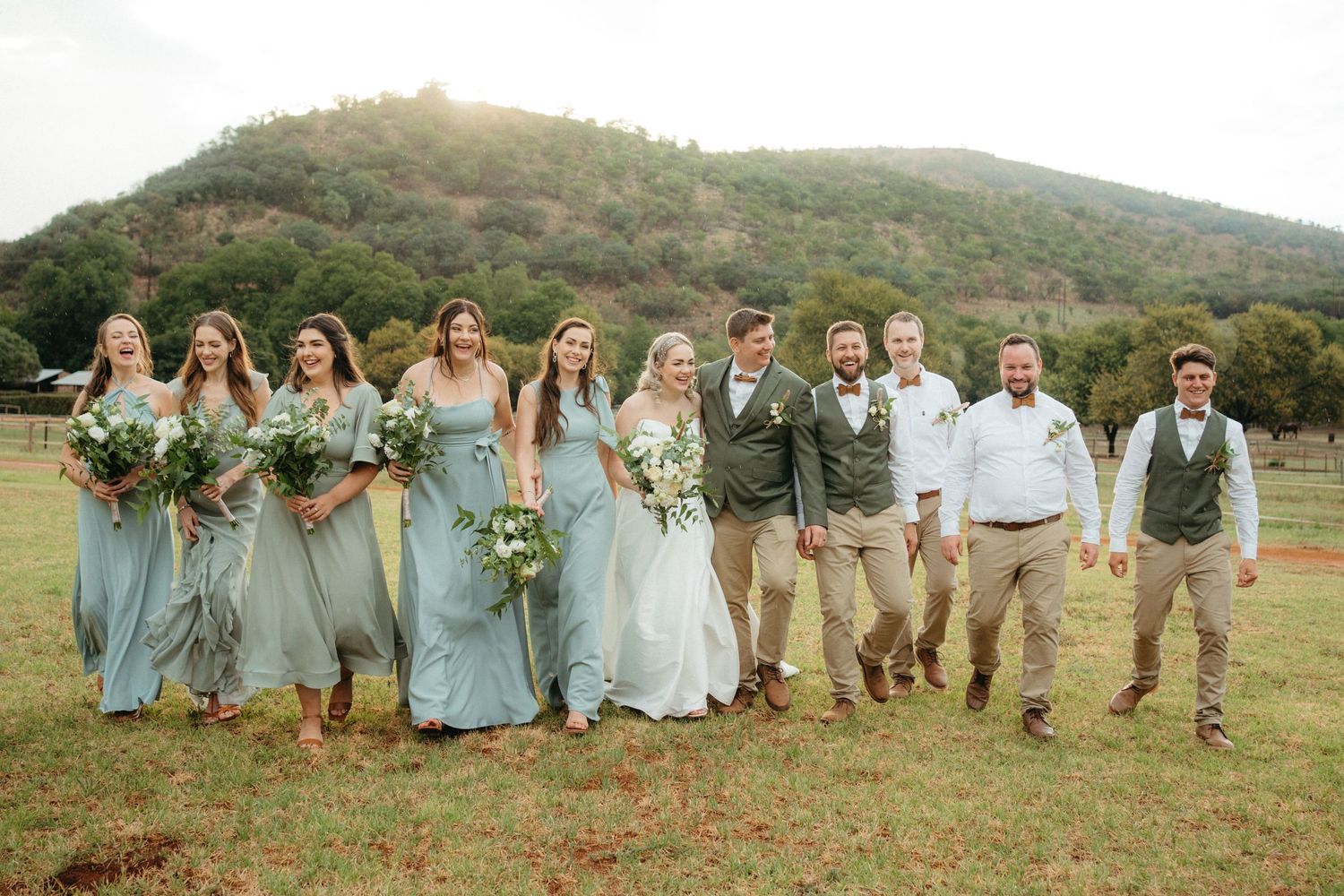 Wedding group poses cheerfully in casual attire against a mountain landscape with dusty blue bridesmaid dresses.