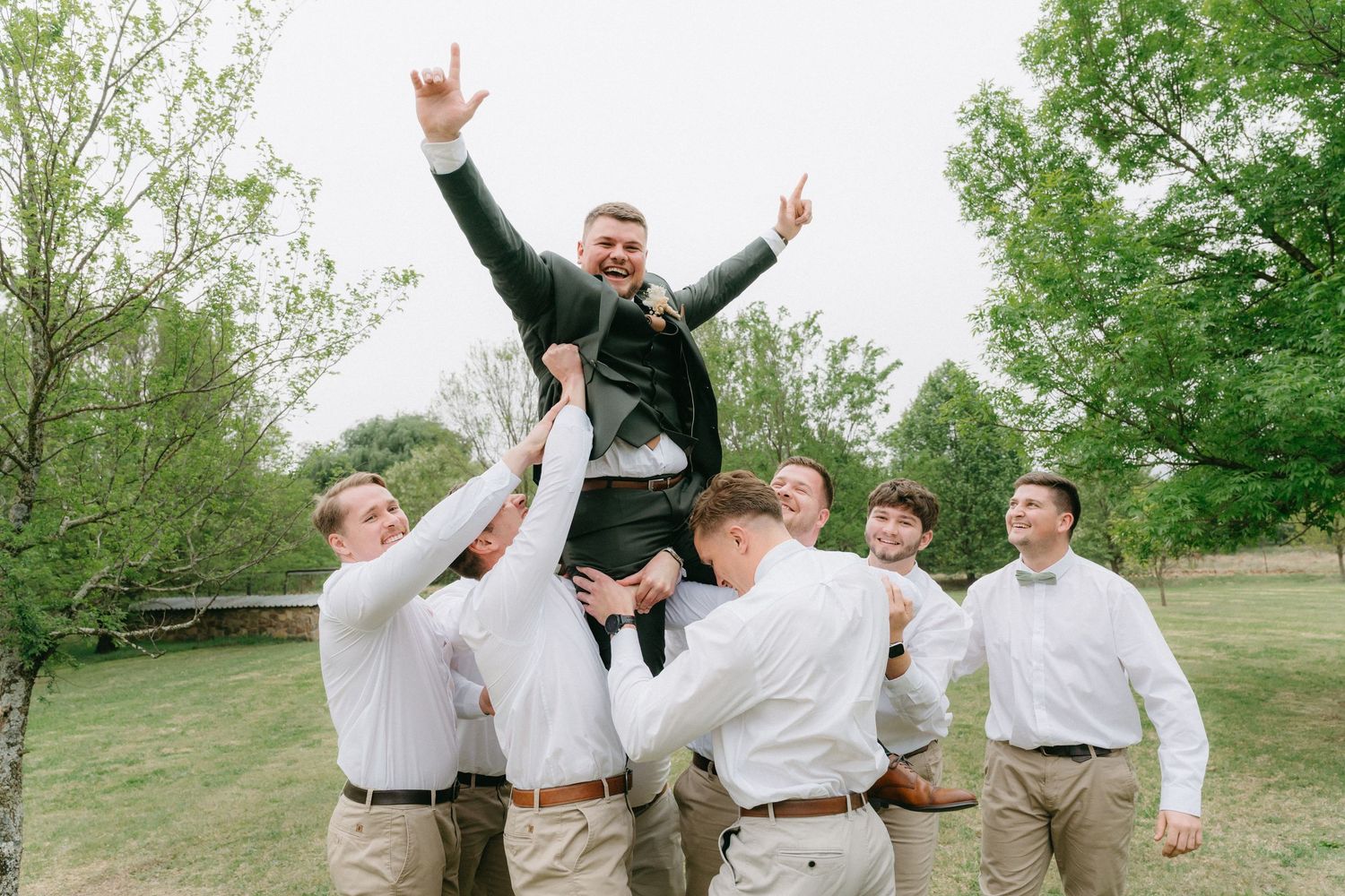 Groomsmen in white shirts celebrate joyfully on grassy hillside.