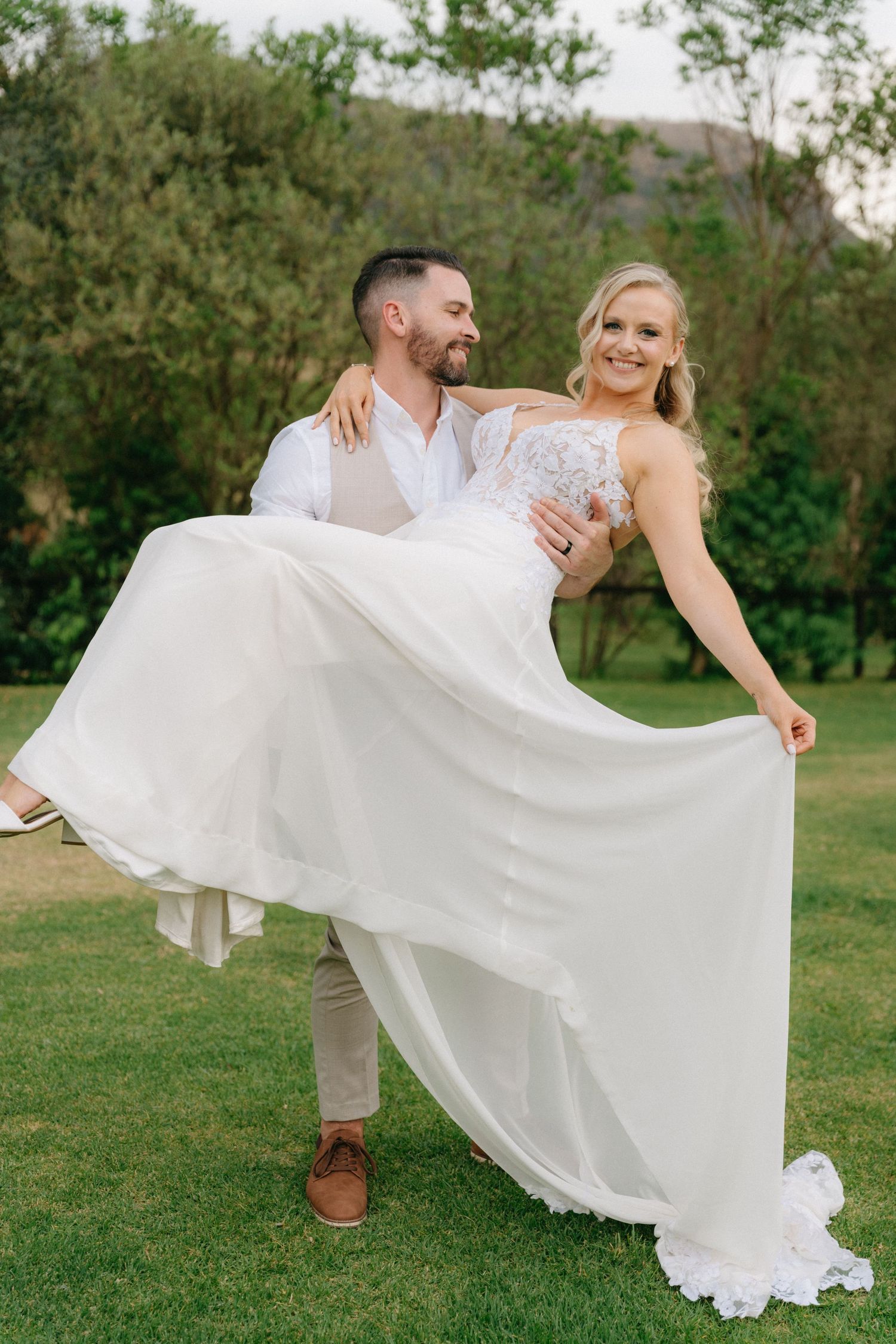 Romantic wedding couple dances playfully on green lawn with flowing white dress billowing in the breeze.