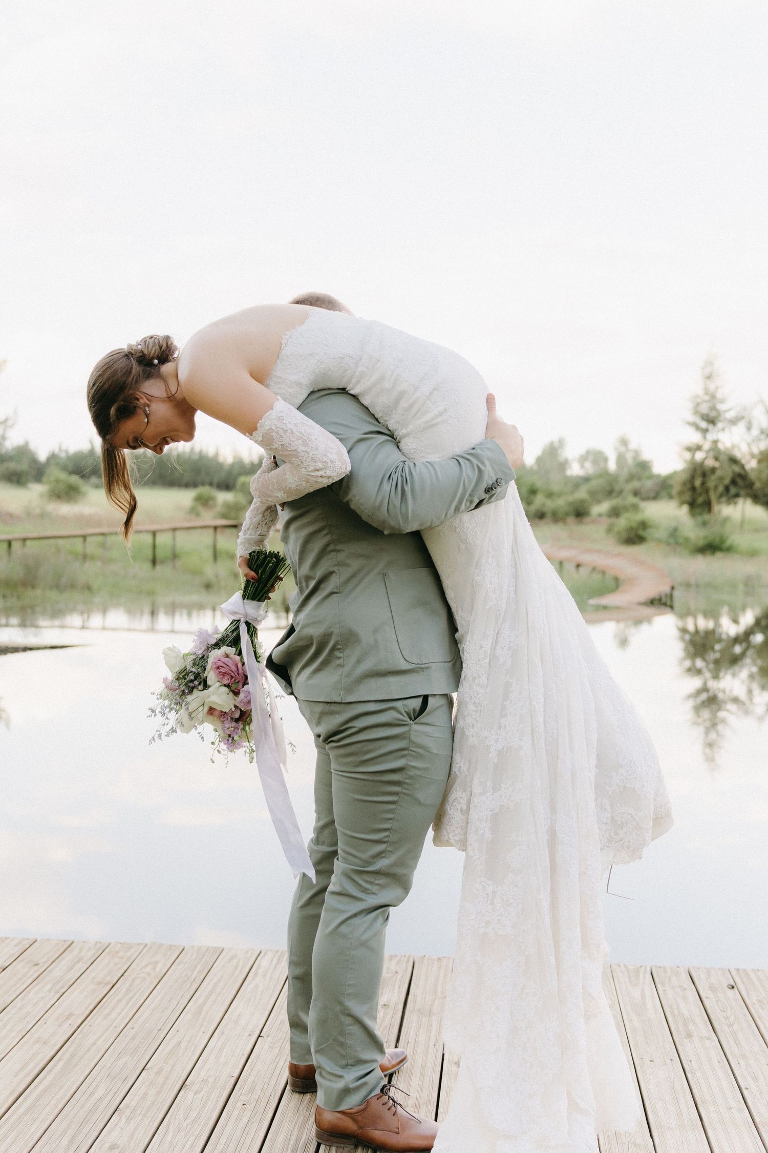 A newlywed couple embraces in a dramatic pose on a wooden dock by a lake.
