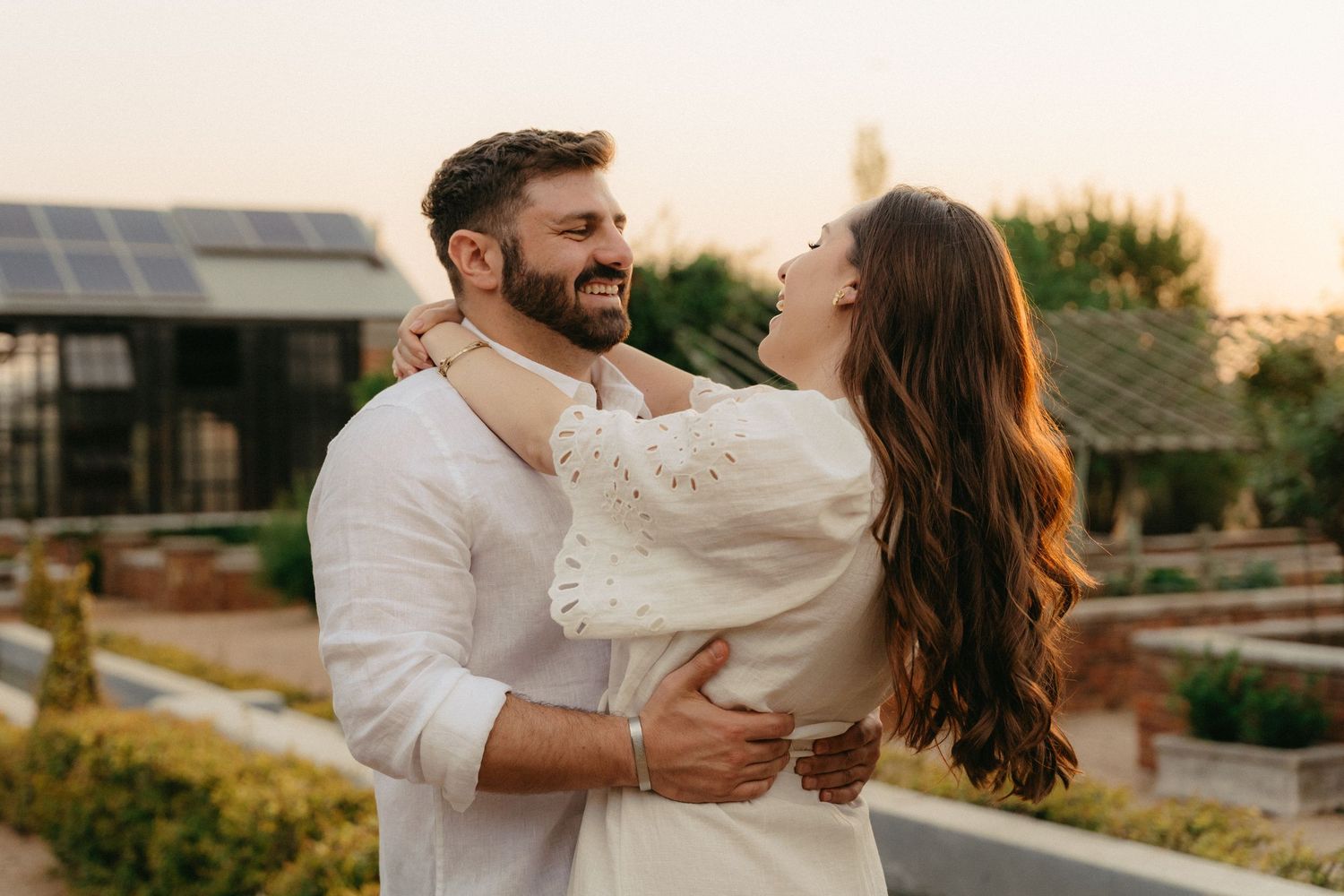A couple in white clothing embraces and laughs together during a romantic sunset photoshoot outdoors.