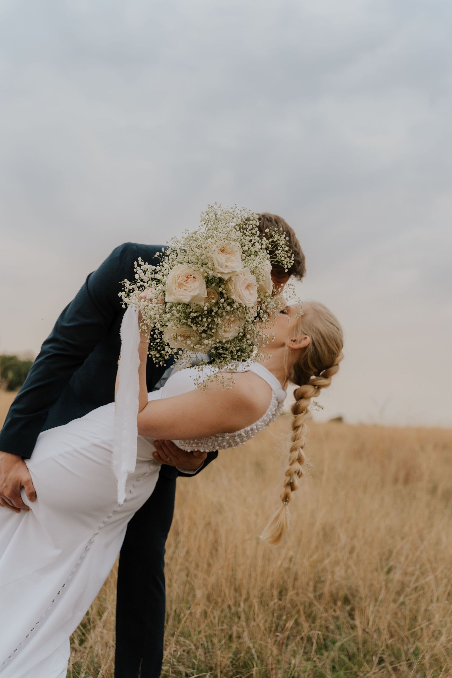 Romantic couple shares a kiss in a golden wheat field at sunset with delicate baby's breath flowers in their hands.