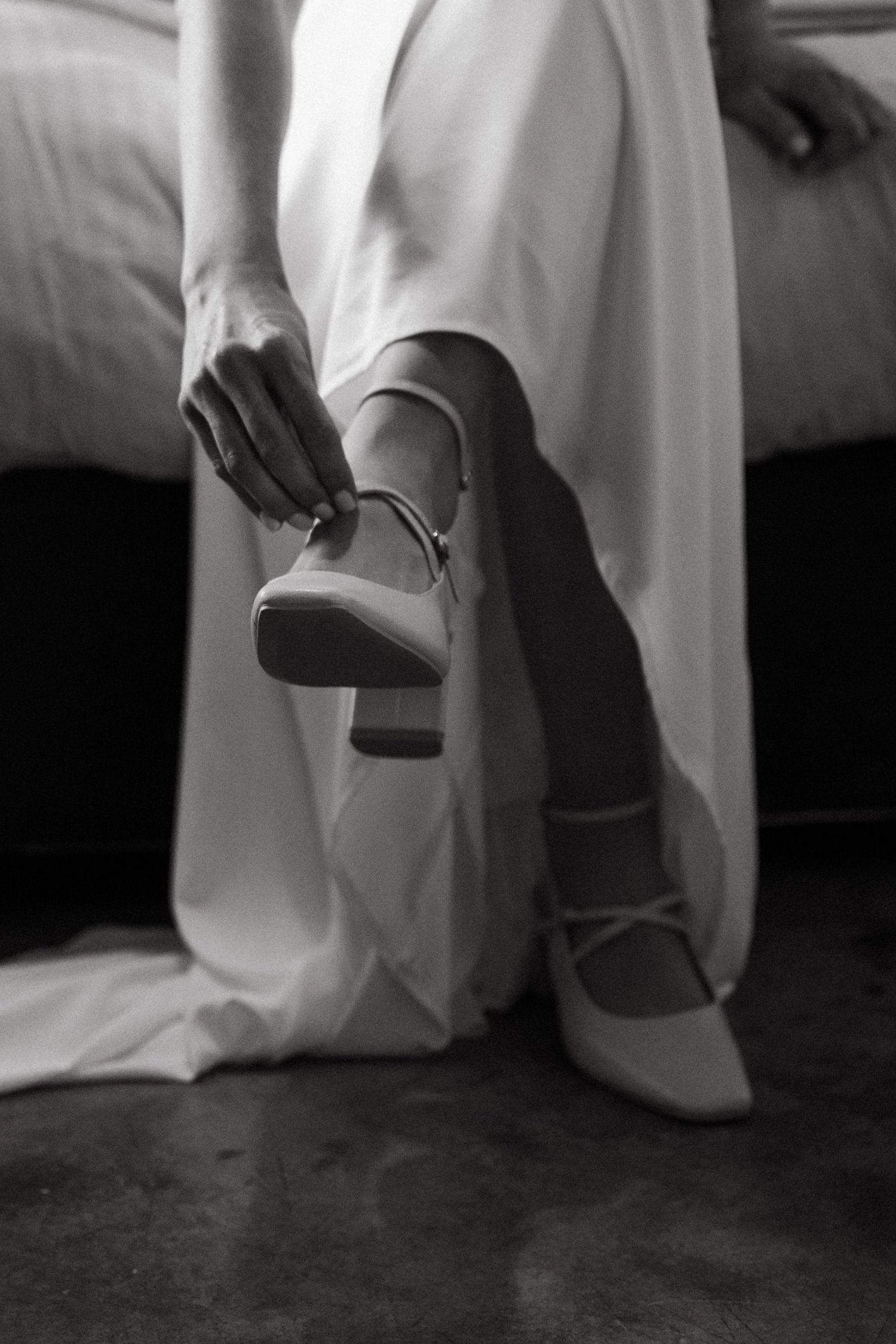 Black and white close-up of bride's shoes and dress hem against dark floor.
