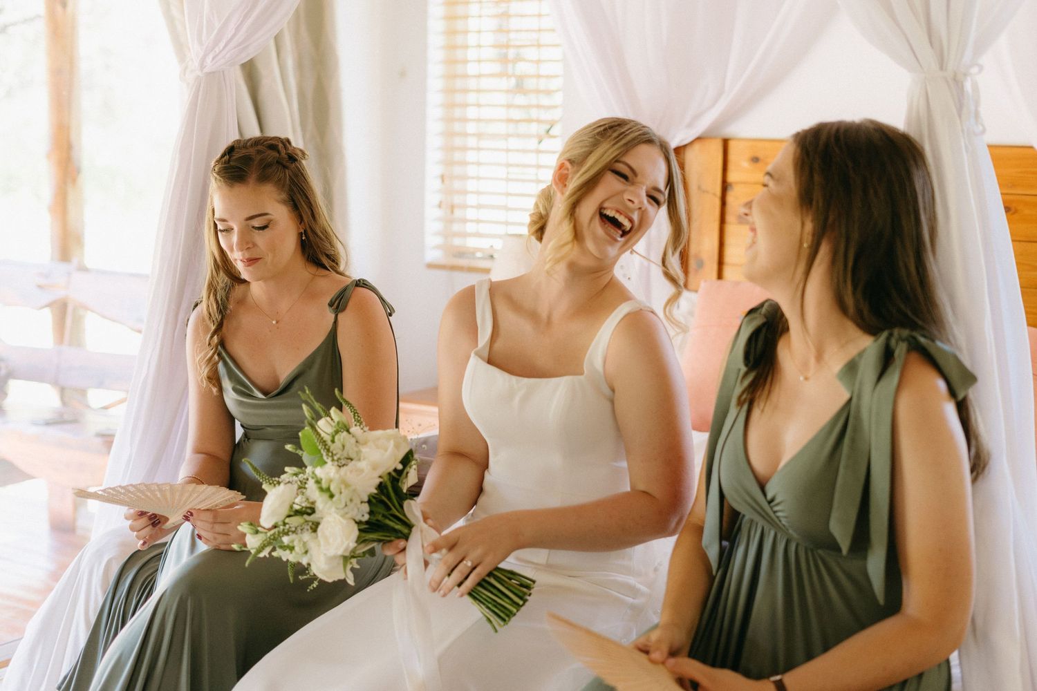 Wedding party sharing joyful moment with bouquet in white draped room.
