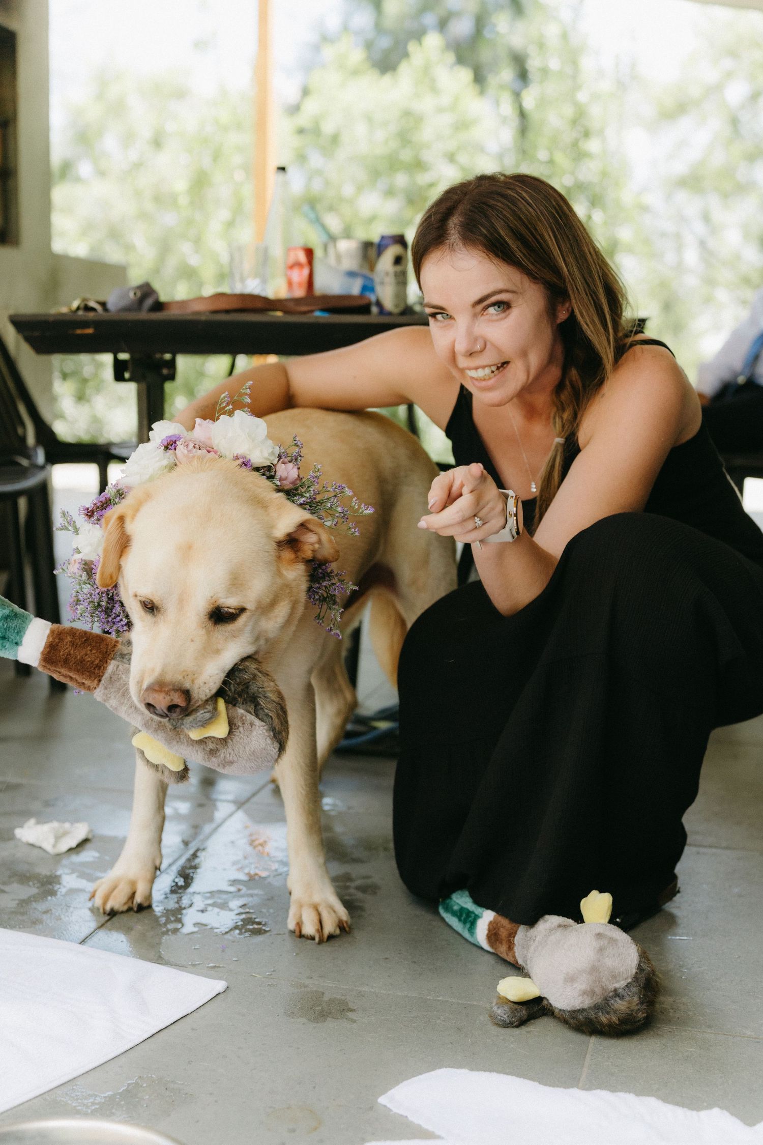 Person in black dress kneels beside yellow Labrador dog wearing flower decorations on its fur.