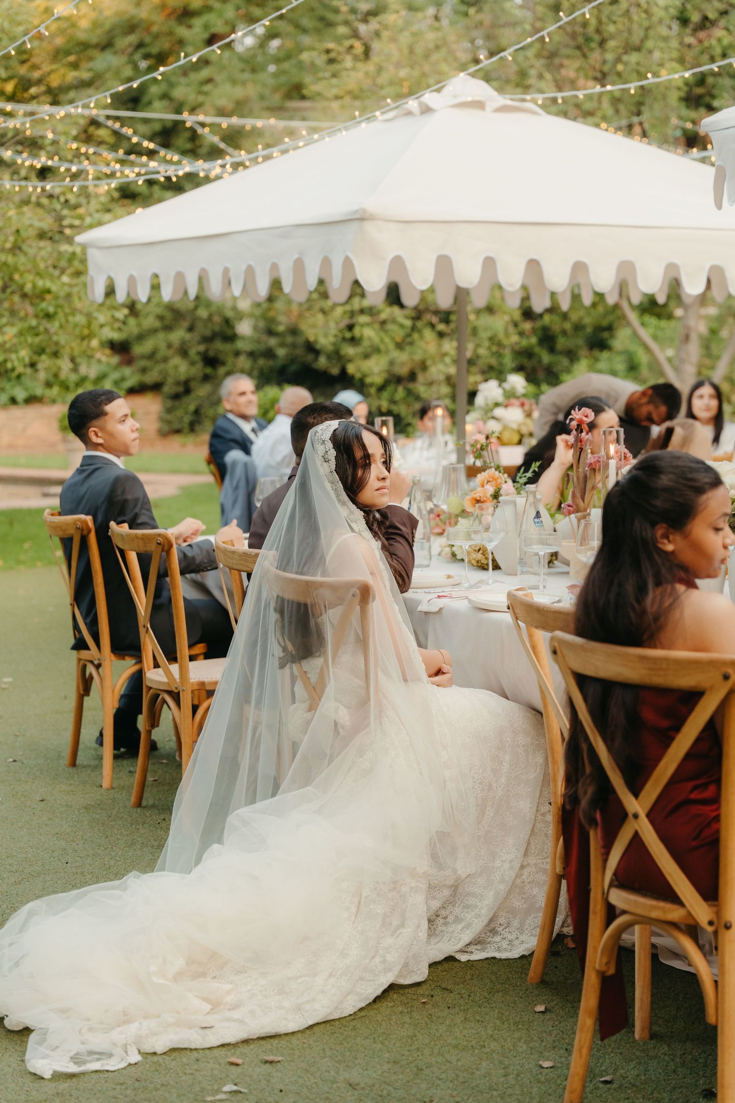Bride in lace gown and veil seated at outdoor wedding reception table under decorative tent with string lights.