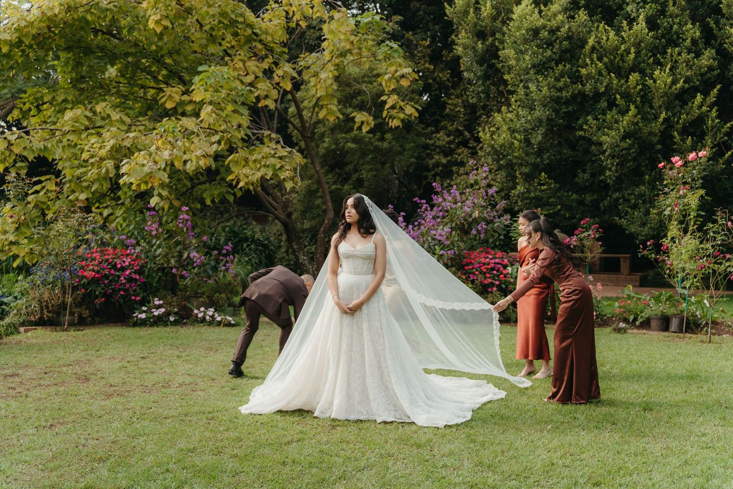 Bride in a white ball gown with veil held by two women in a lush garden with colorful flowers and trees.