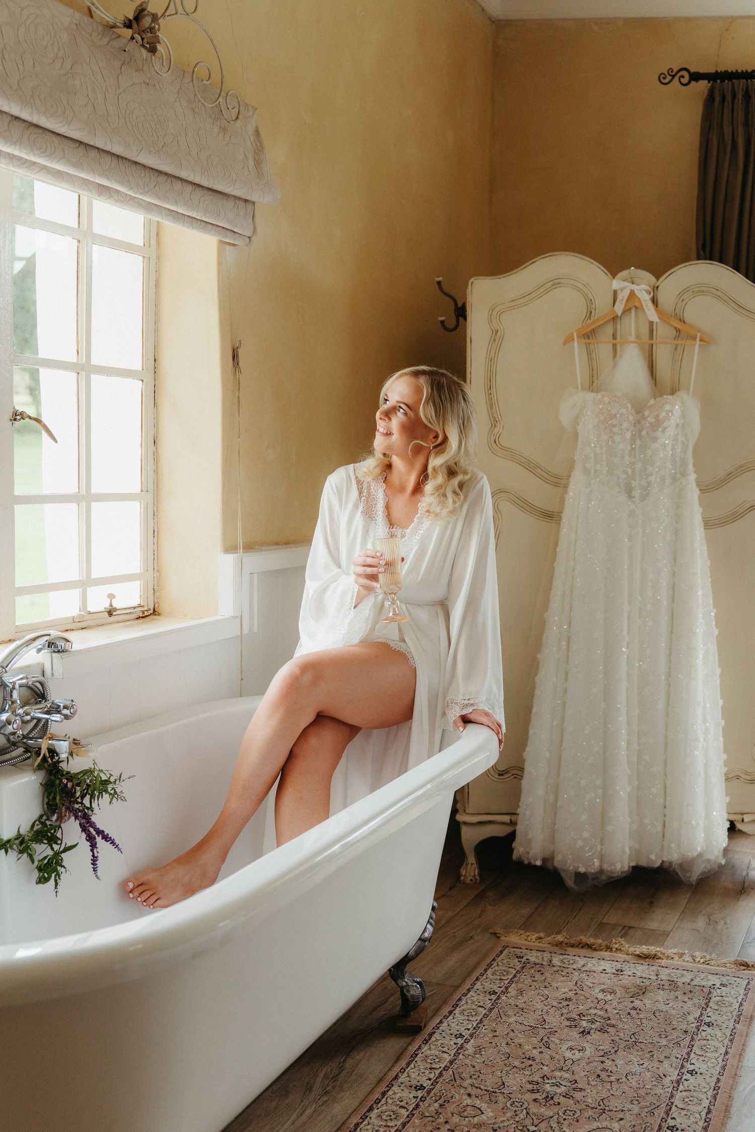 Bride in white robe sitting on clawfoot bathtub holding champagne glass with wedding dress hanging nearby.