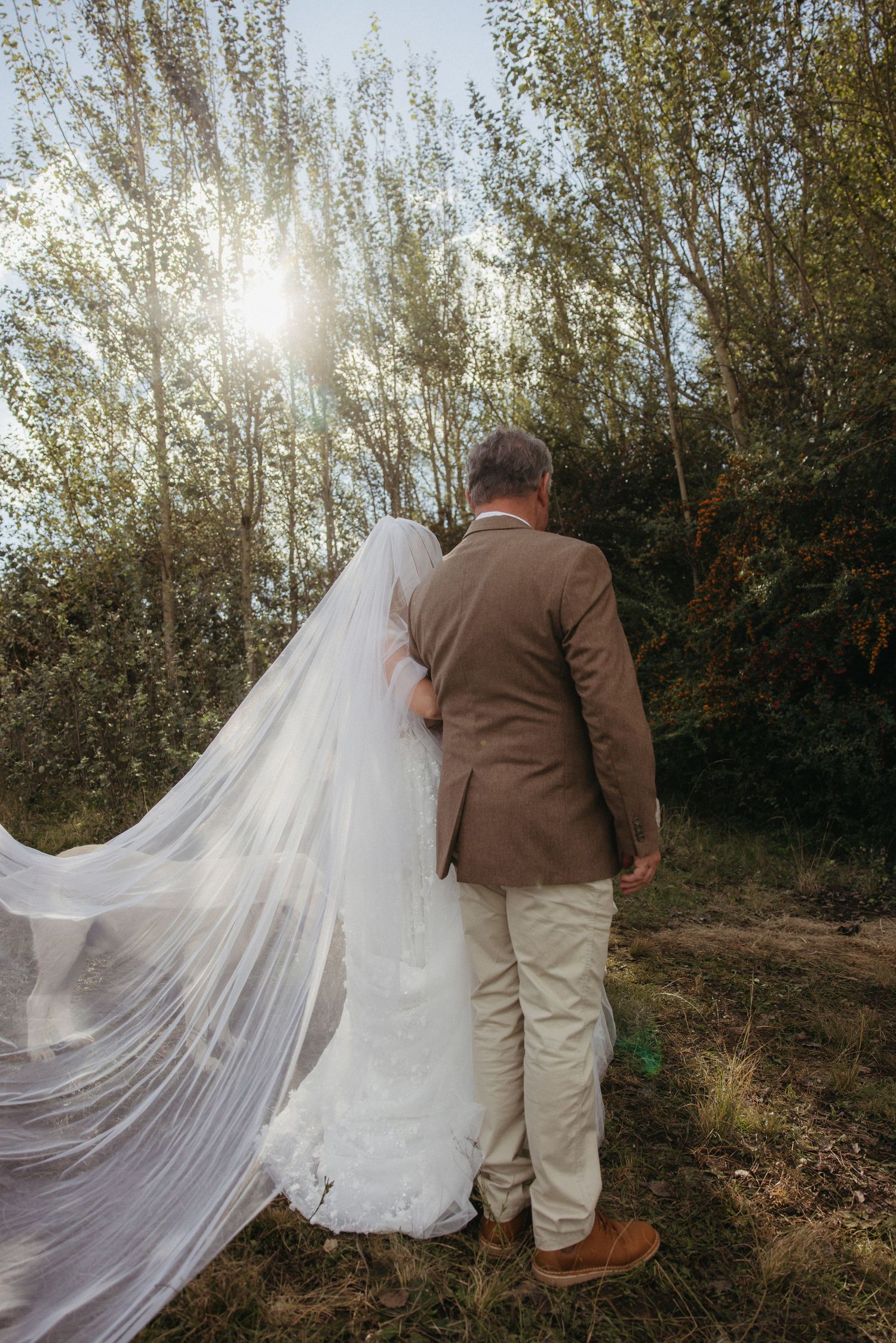 Bride in flowing white veil and groom in tan suit walk hand in hand through sunlit forest on wedding day.