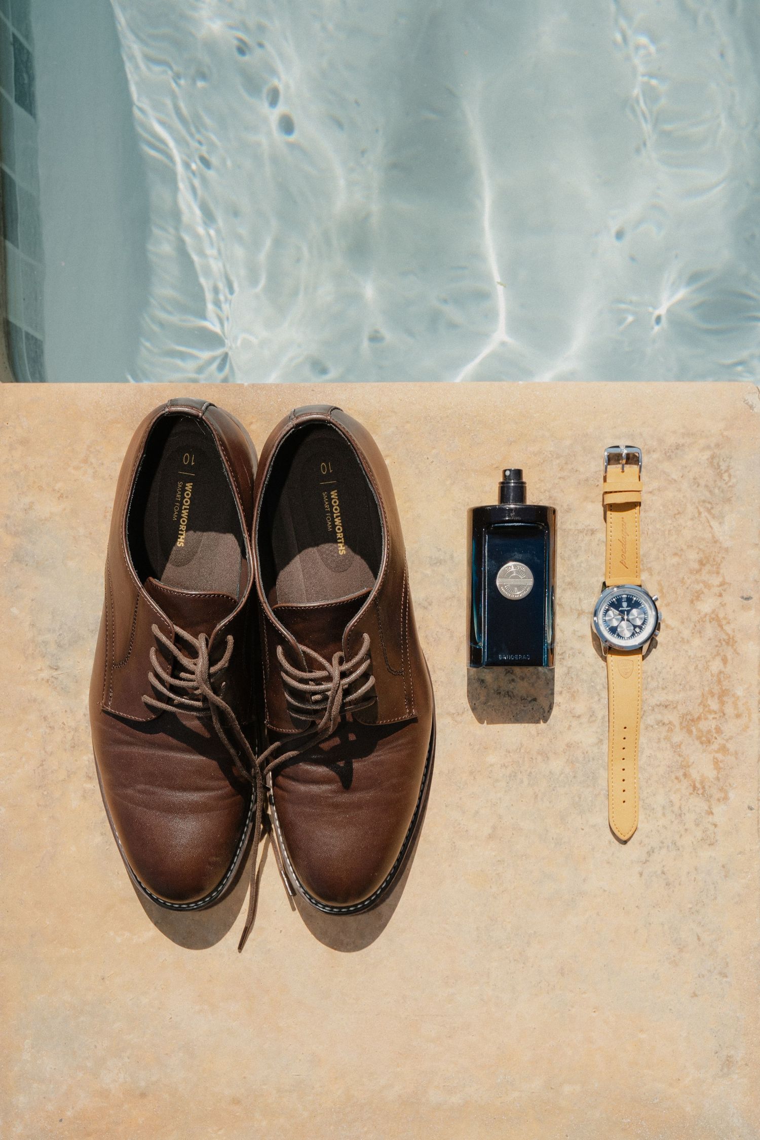 Brown leather dress shoes, dark cologne bottle, and tan-strapped watch displayed on stone surface near pool.