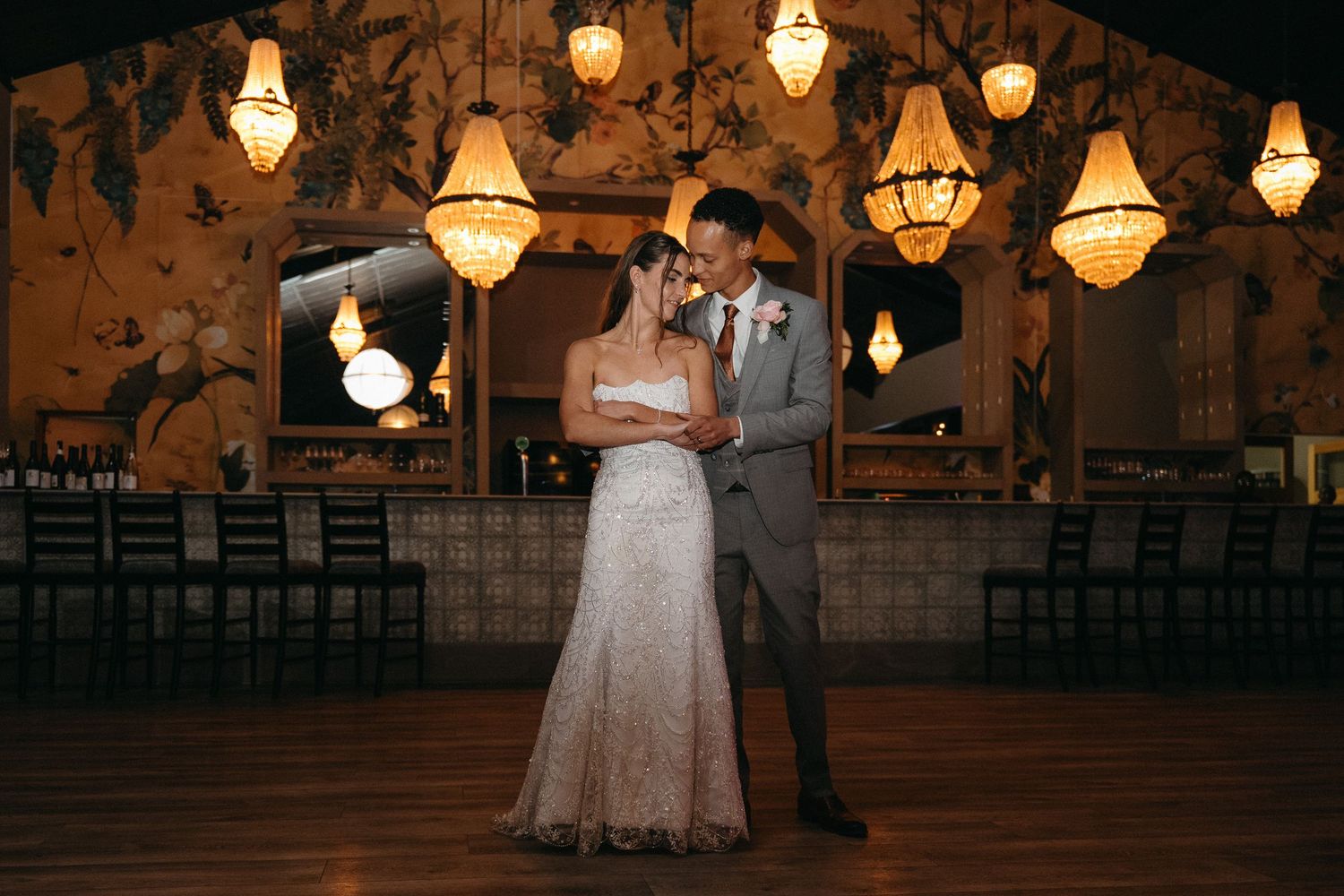 Bride and groom share first dance under elegant chandeliers in a warmly lit wedding reception venue.
