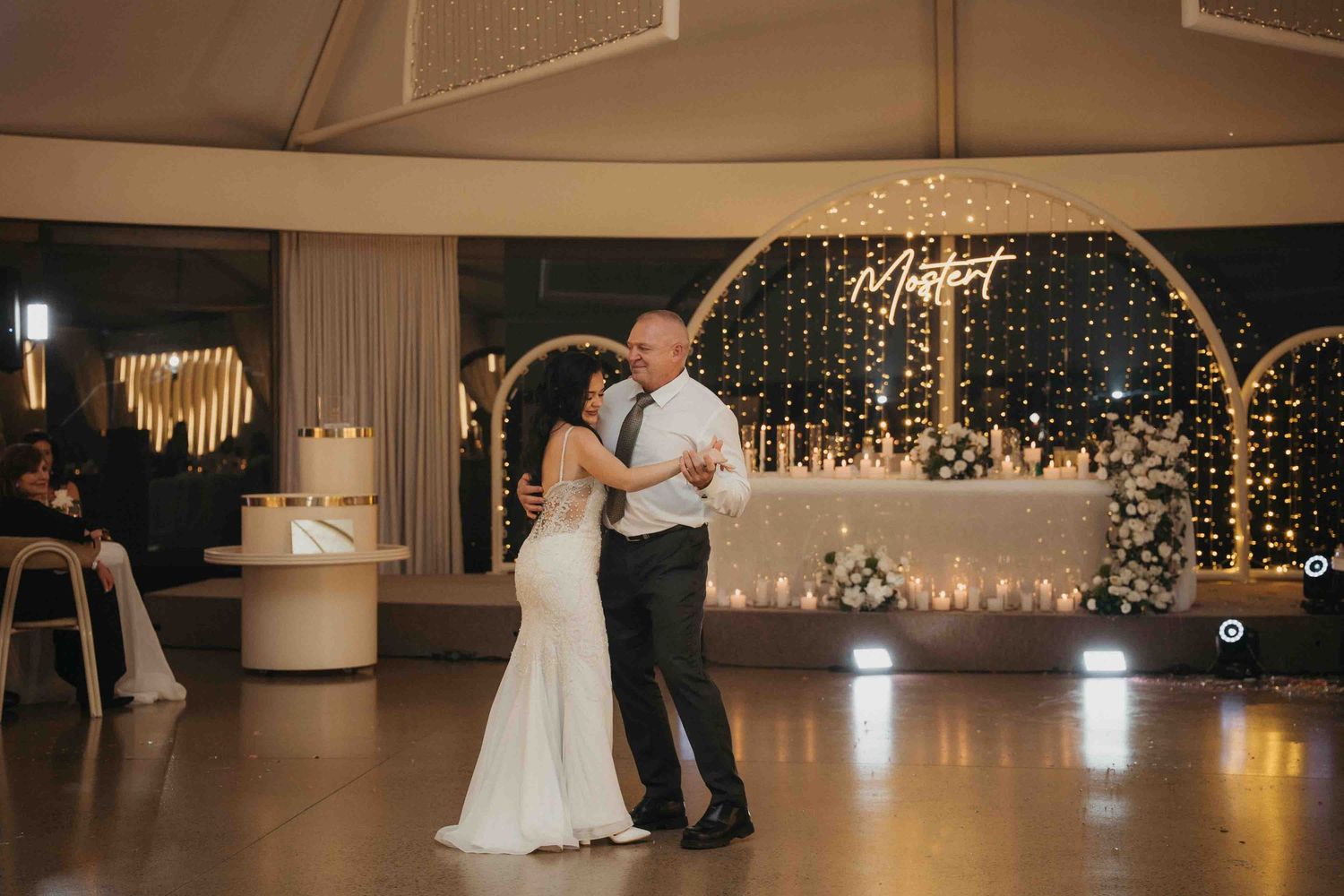 Bride and groom share their first dance in an elegant wedding venue with warm fairy lights and floral backdrop.