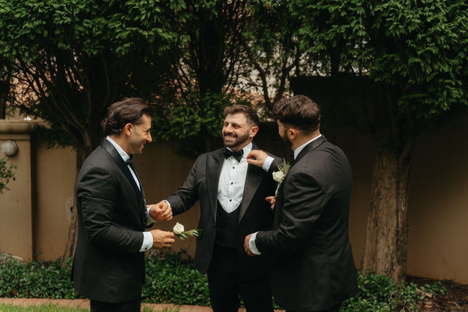 Three groomsmen in black tuxedos laughing and adjusting boutonnieres outdoors before a wedding ceremony.