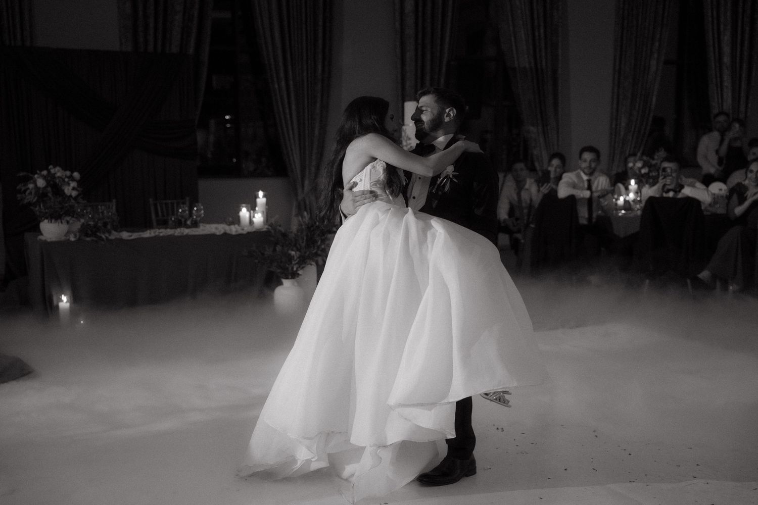 Black and white photo of a bride and groom sharing their first dance, surrounded by guests at an elegant wedding reception.