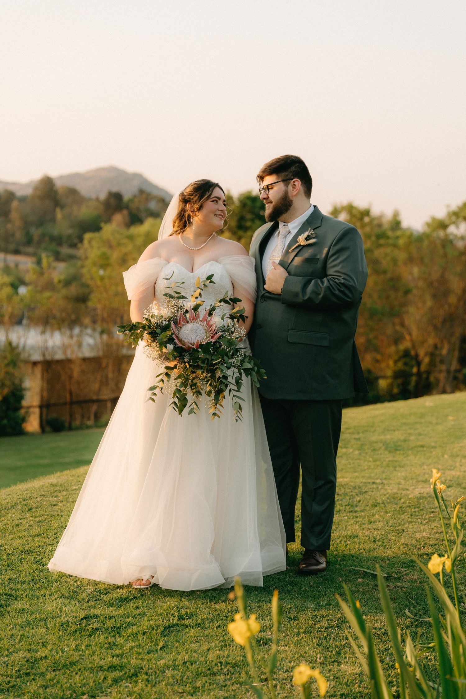 Bride and groom standing on a green lawn at sunset, at Cradle Valley Lodge with Muldersdrift mountains and a bouquet in the background.