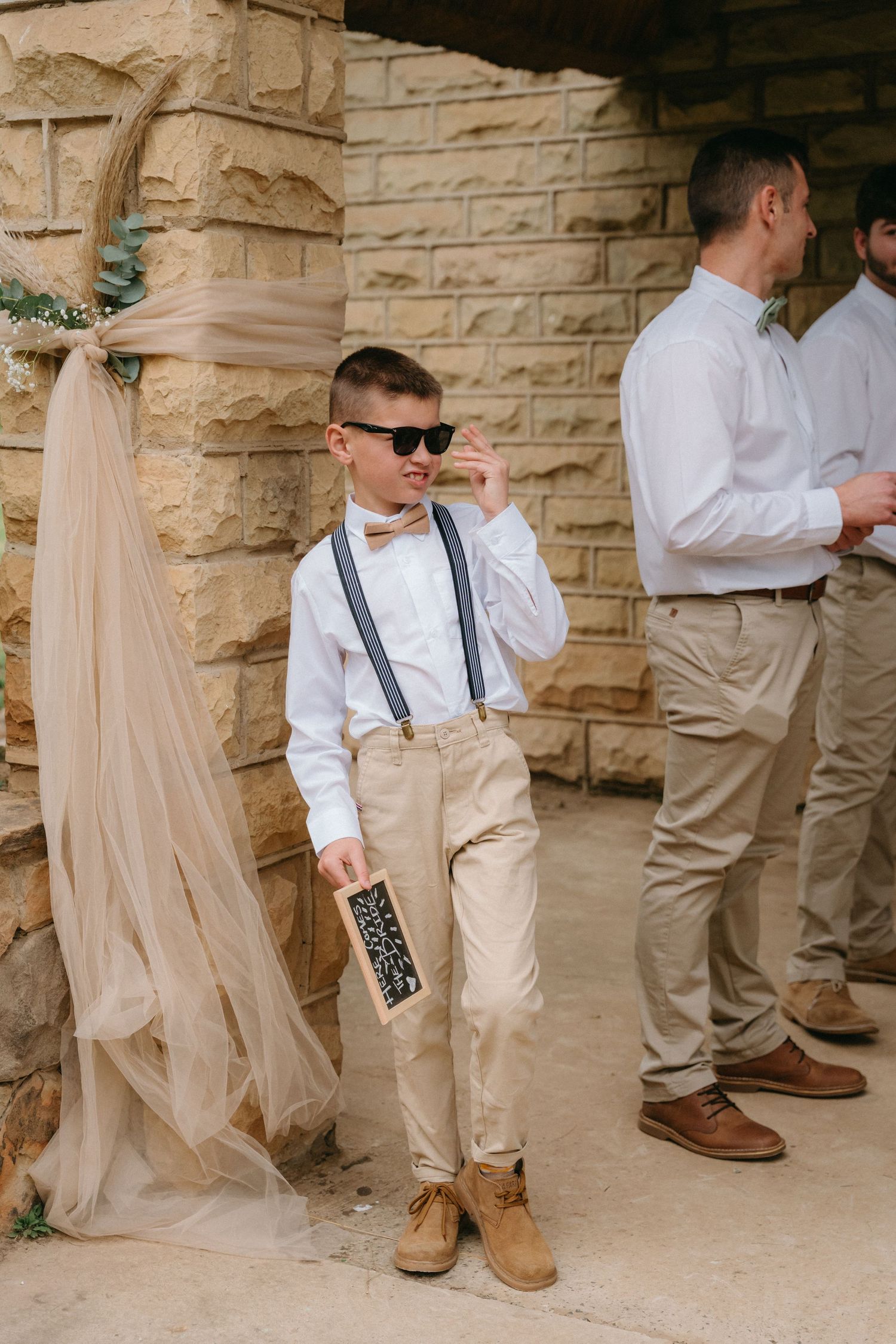 Groomsmen in white shirts and suspenders stand against stone wall showing details of wedding attire.