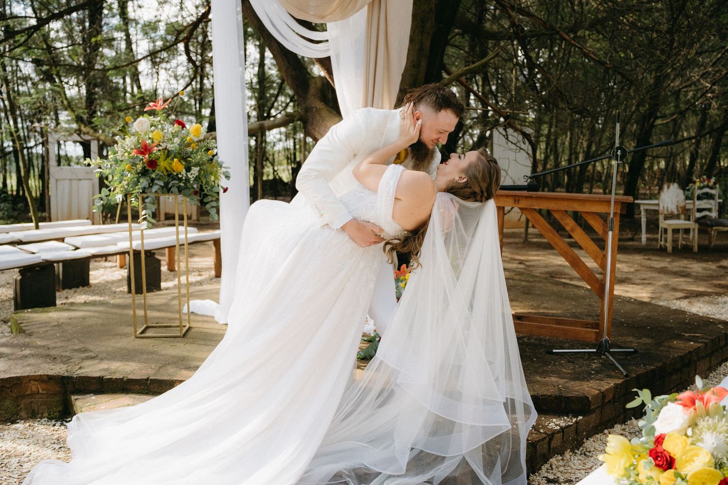 A romantic wedding ceremony moment under a draped canopy in a wooded outdoor setting with floral decorations.