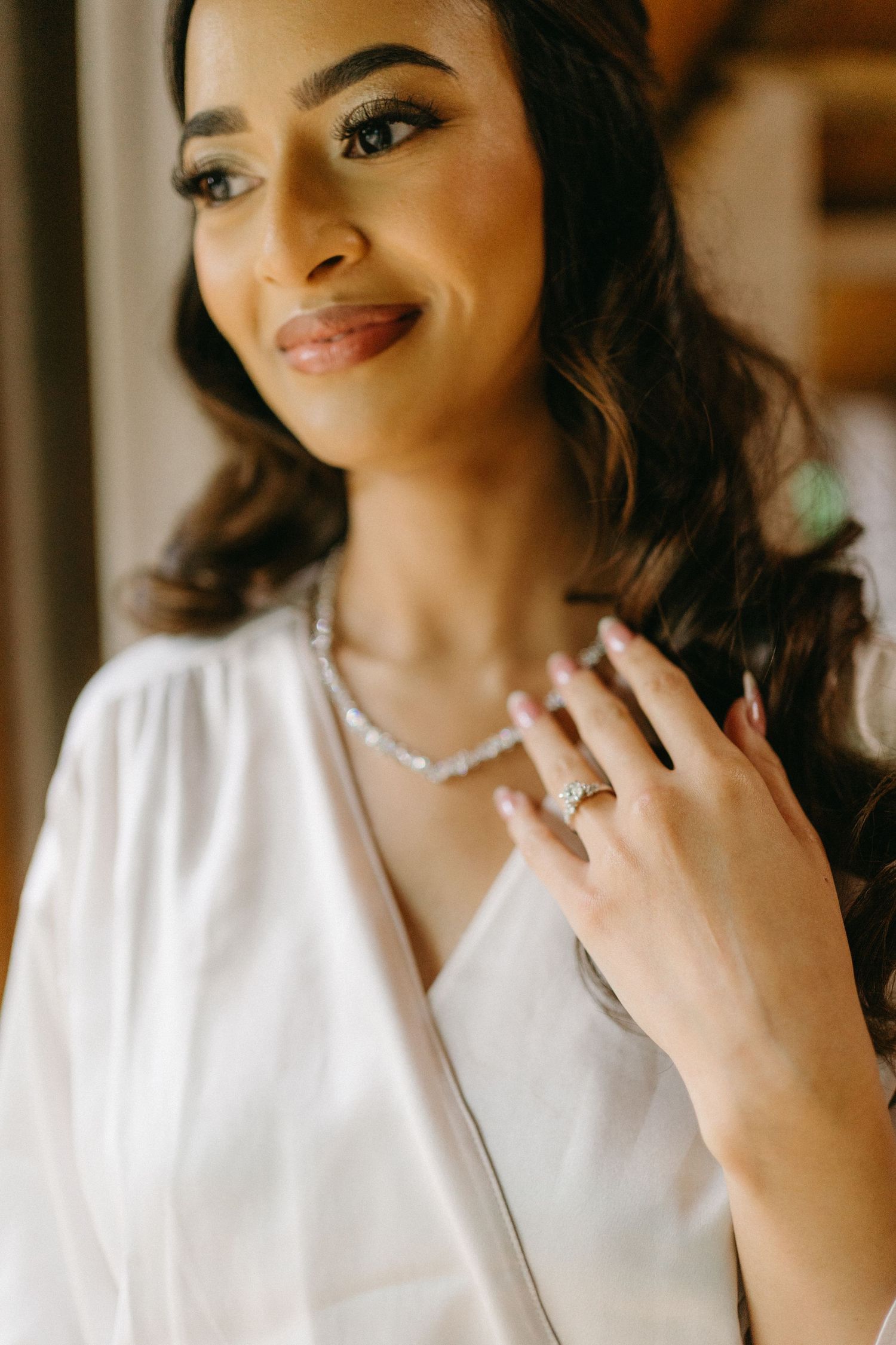 Woman in white robe wearing diamond necklace and ring poses elegantly by window.