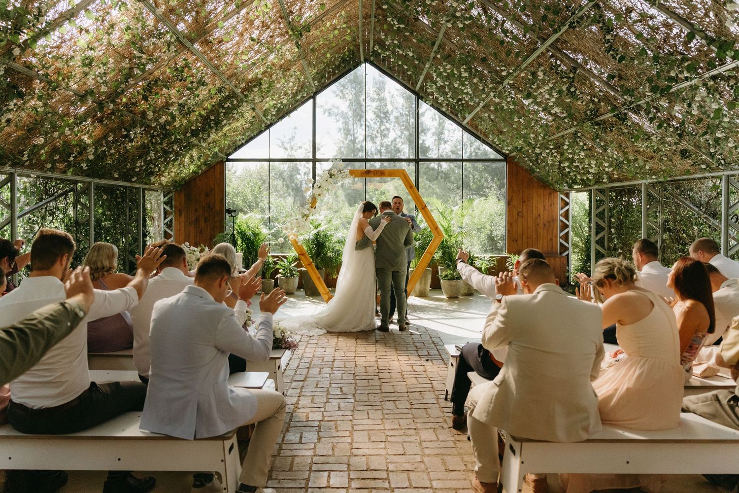 Wedding ceremony taking place in a sunlit greenhouse with guests seated on wooden benches.