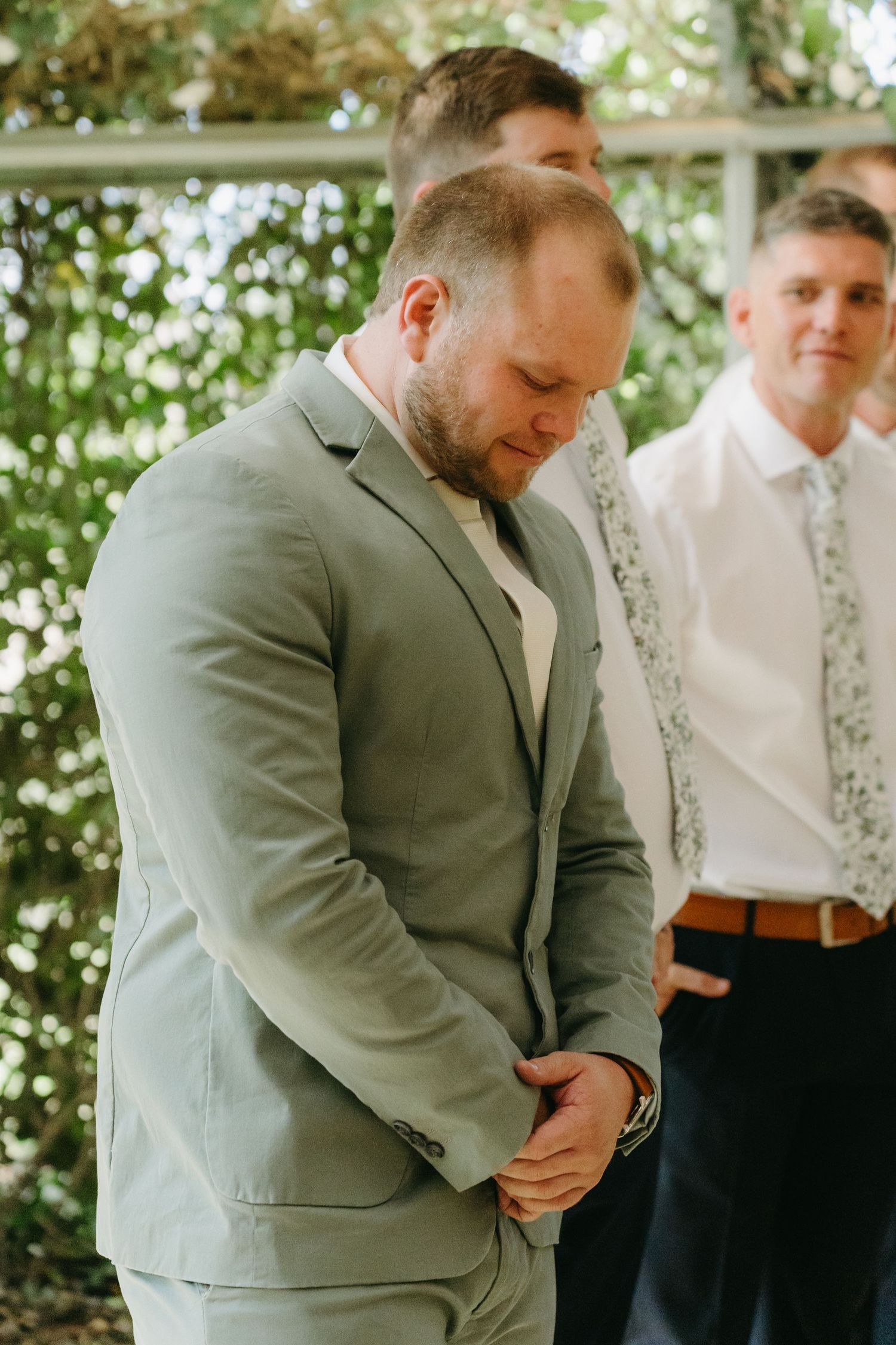 A groom in a light gray suit bows his head during an emotional moment at an outdoor wedding ceremony.