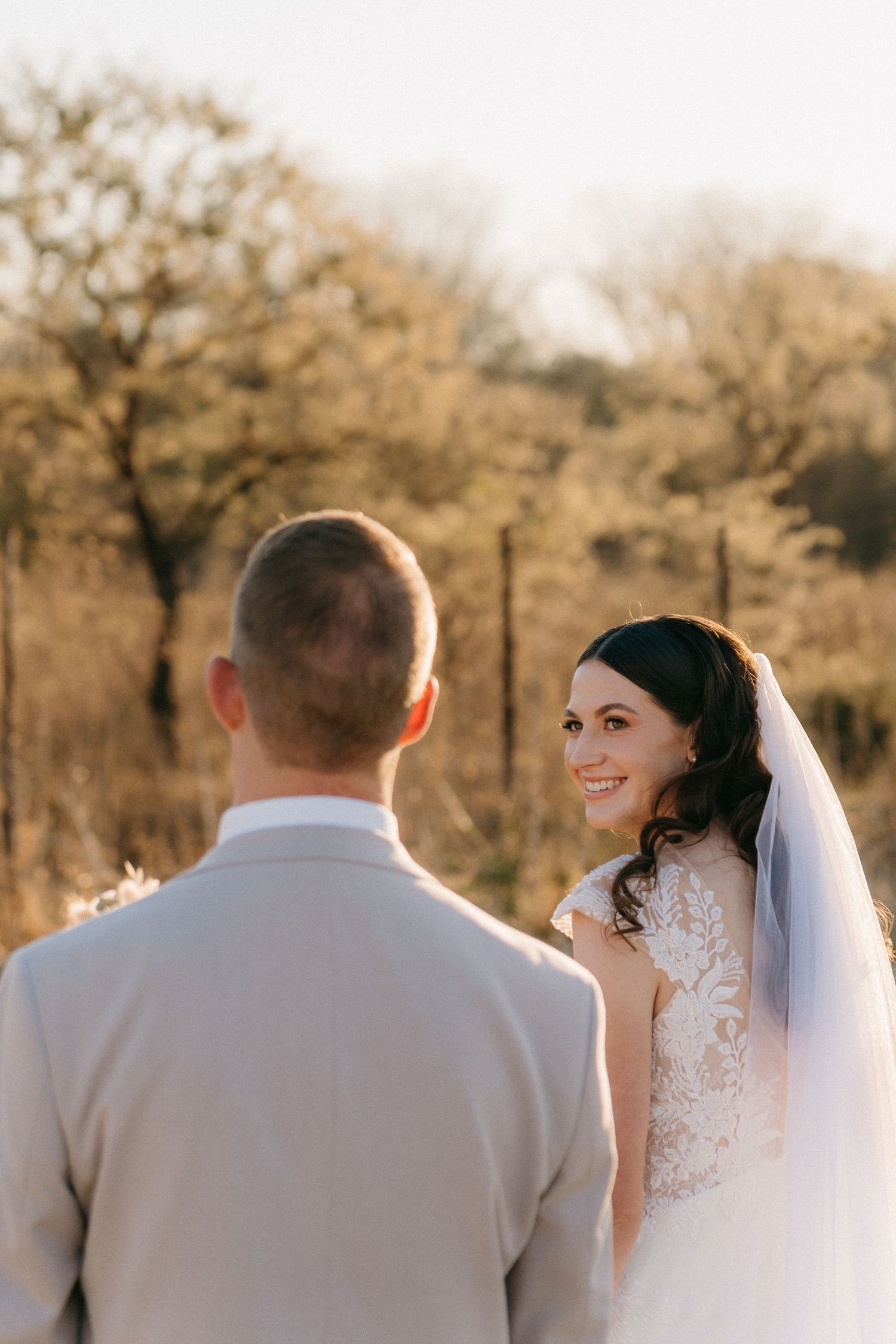 A bride in a lace wedding dress smiles over her shoulder during a golden hour outdoor photoshoot.