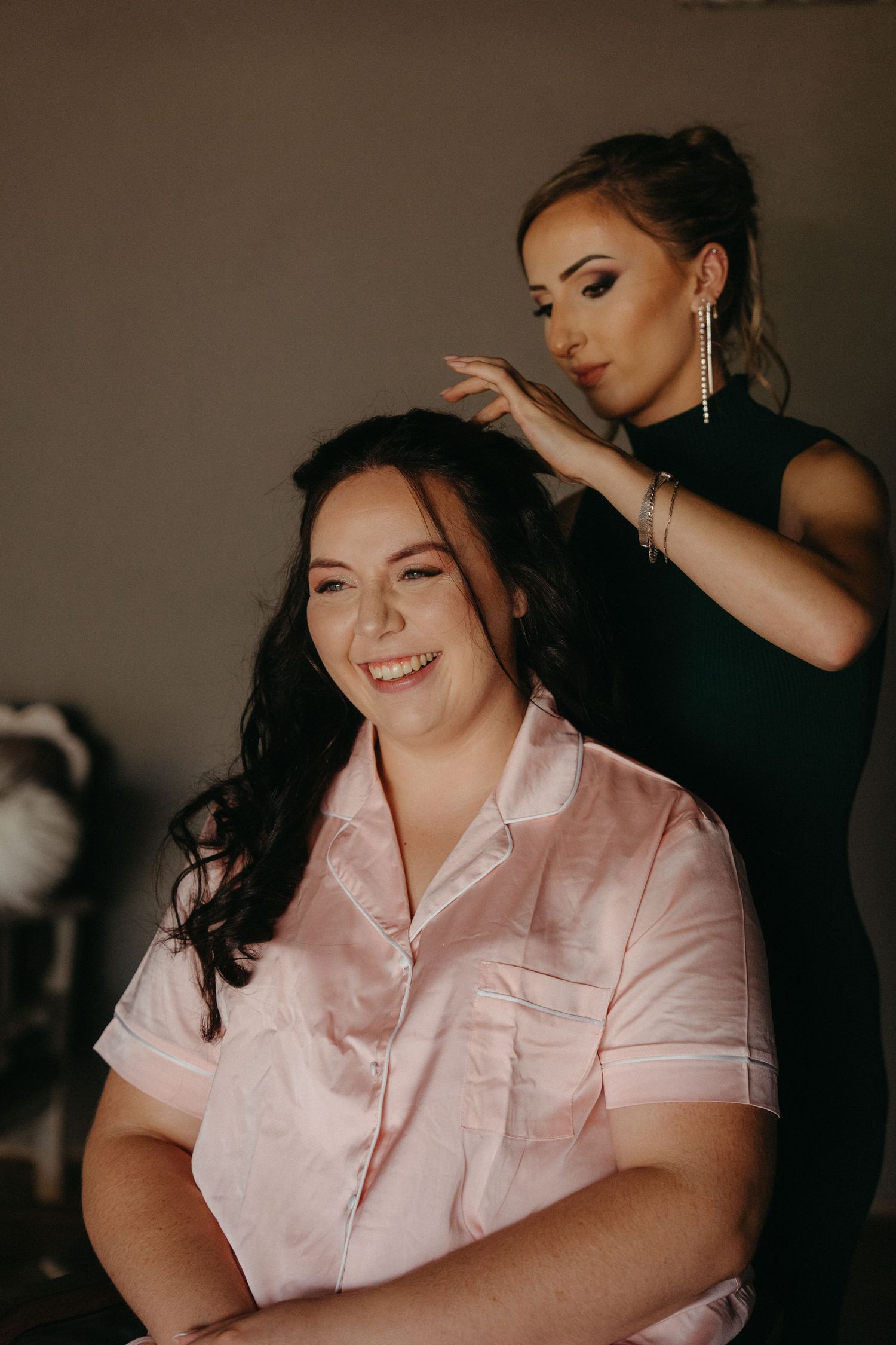 A stylist working on a client's hair while she smiles warmly in a pink satin robe during wedding preparations.