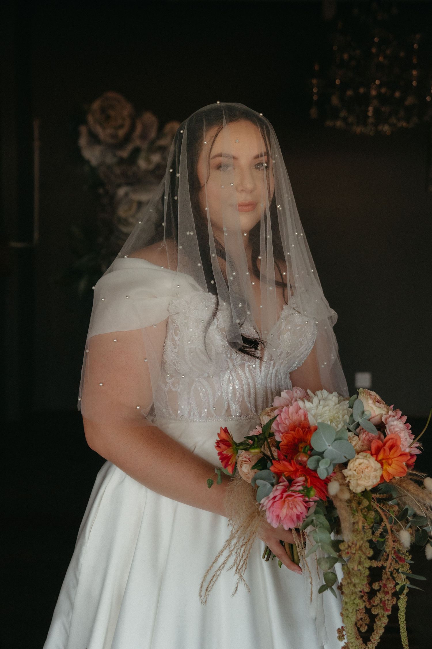 A bride in a white off-shoulder wedding gown and veil holds a vibrant bouquet of pink, orange and coral flowers.