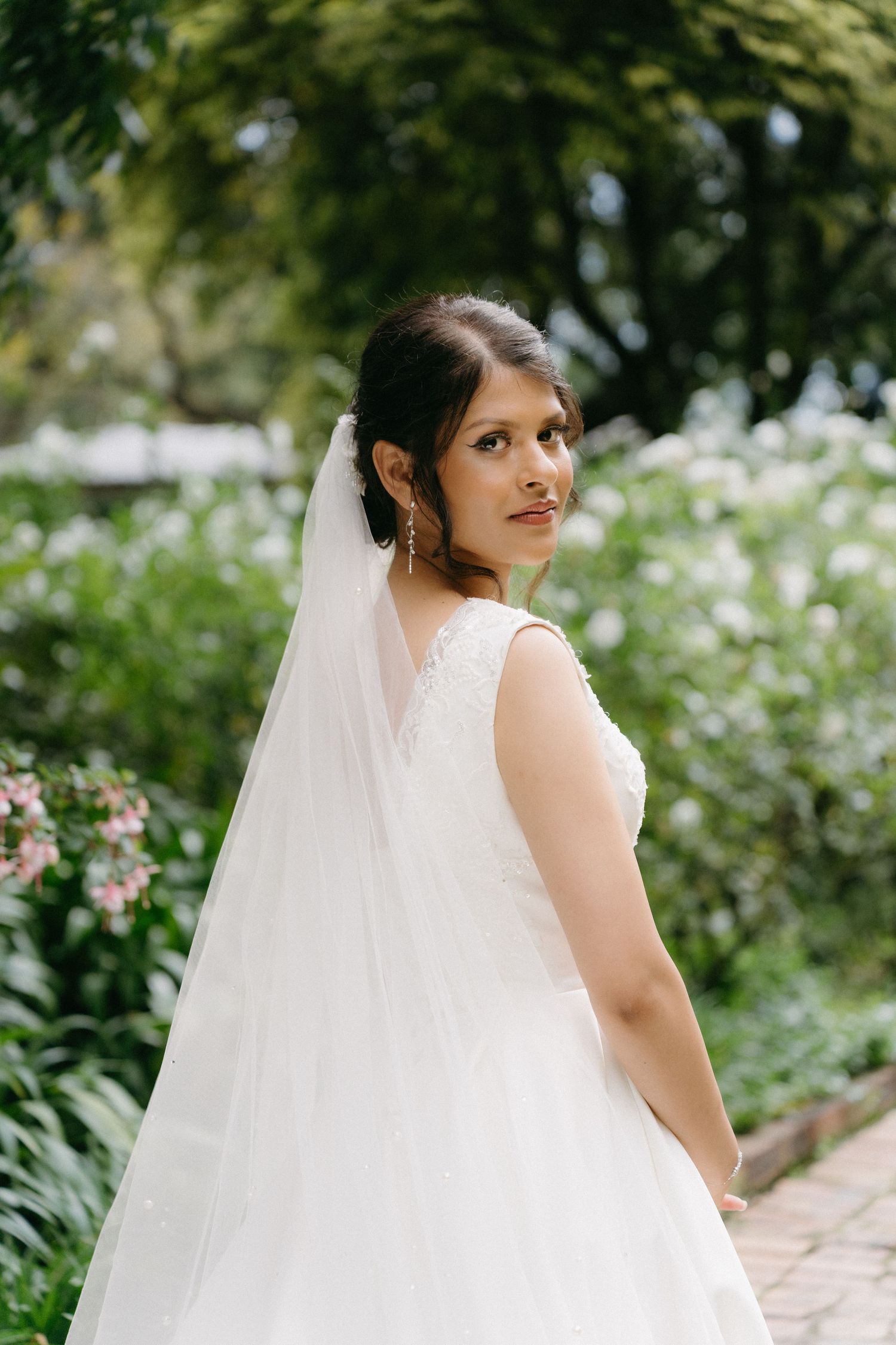 A bride in a classic white wedding dress poses in a garden surrounded by blooming flowers.