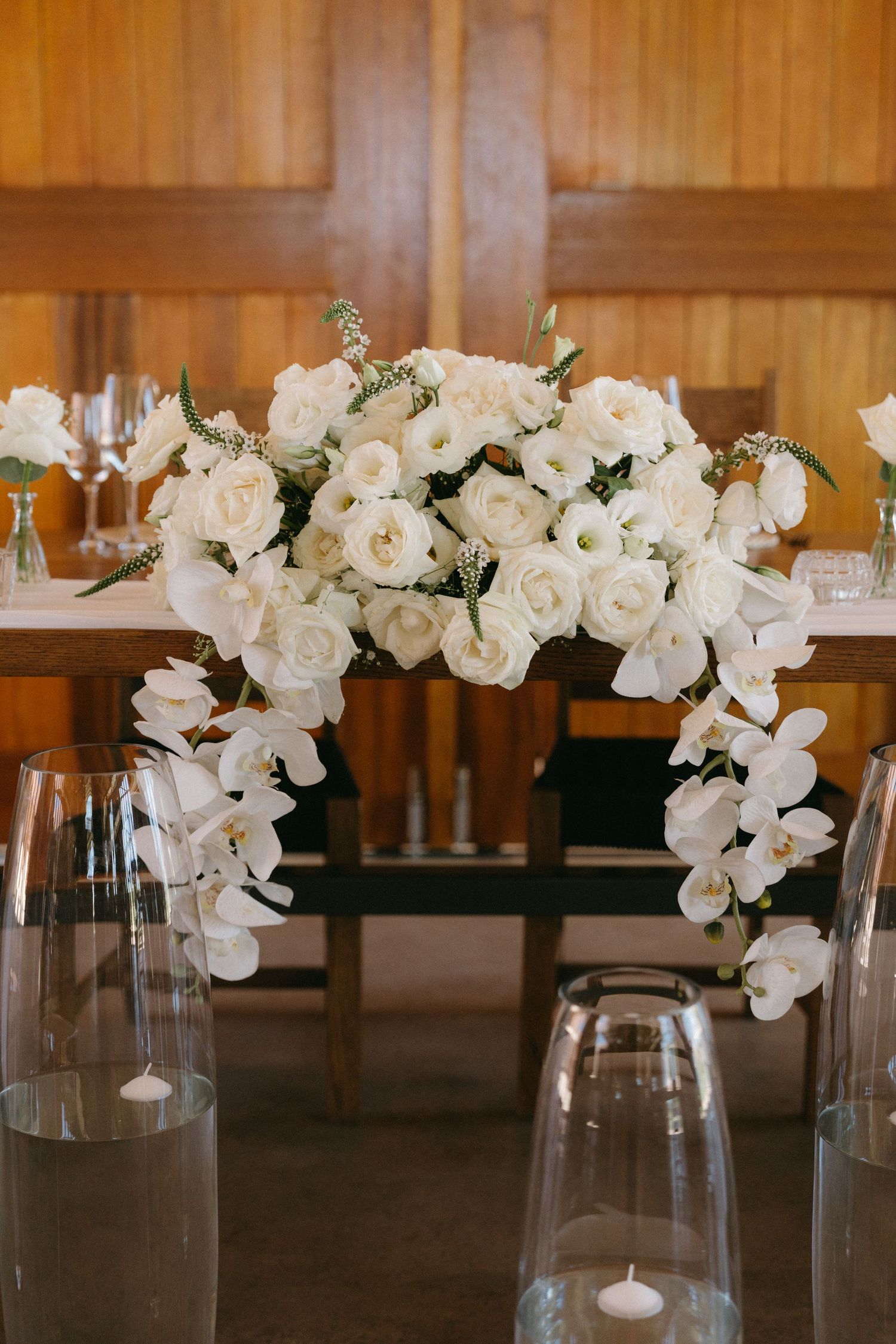 Elegant white rose and orchid floral centerpiece with glass candleholders on wooden table.