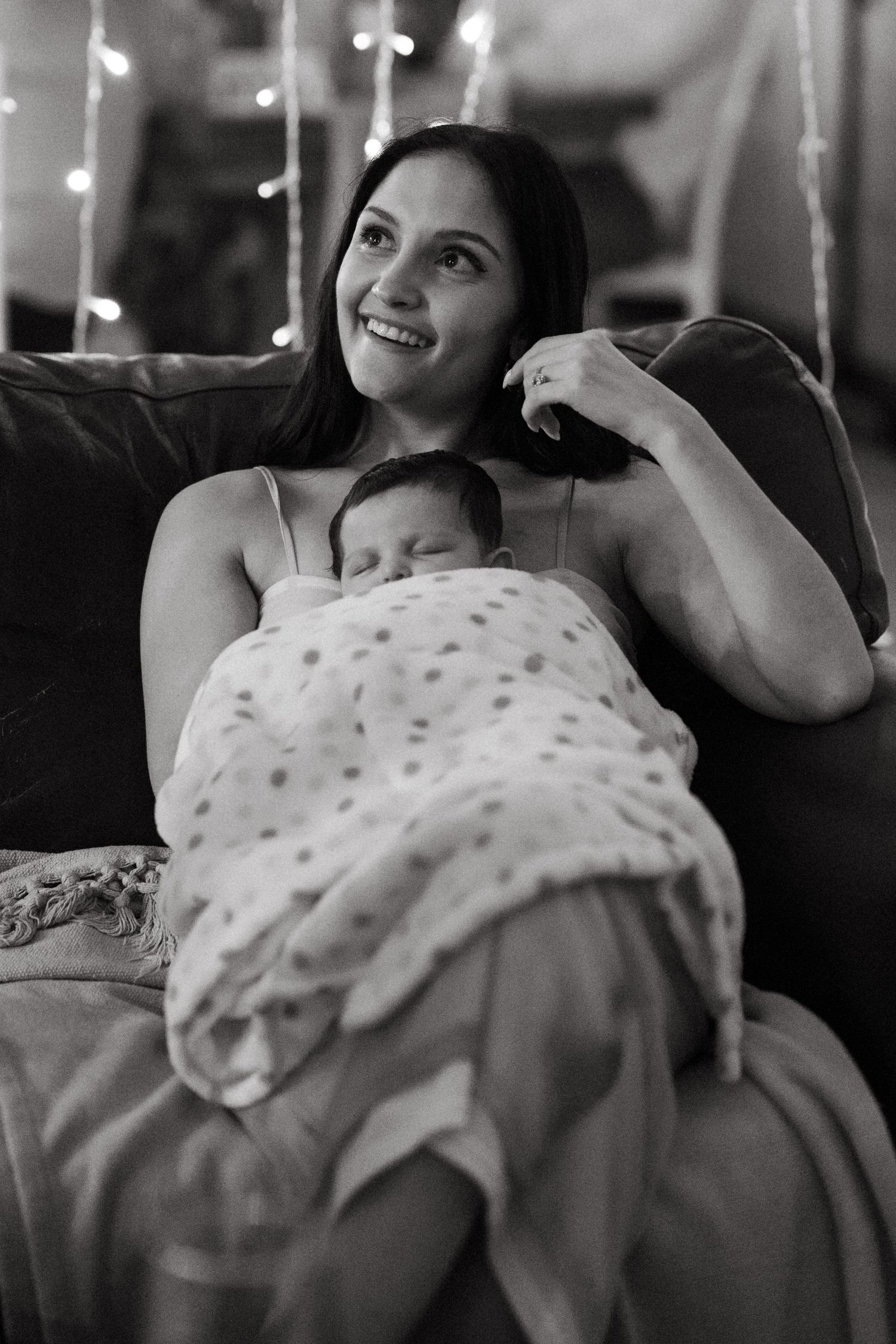 A tender moment captured in black and white as a mother relaxes on a couch with a sleeping baby under twinkling lights.