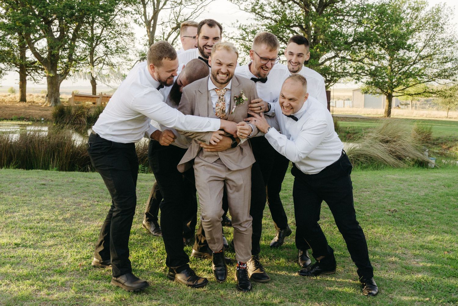 Groomsmen share laughs and embrace on grassy lawn during wedding celebration.