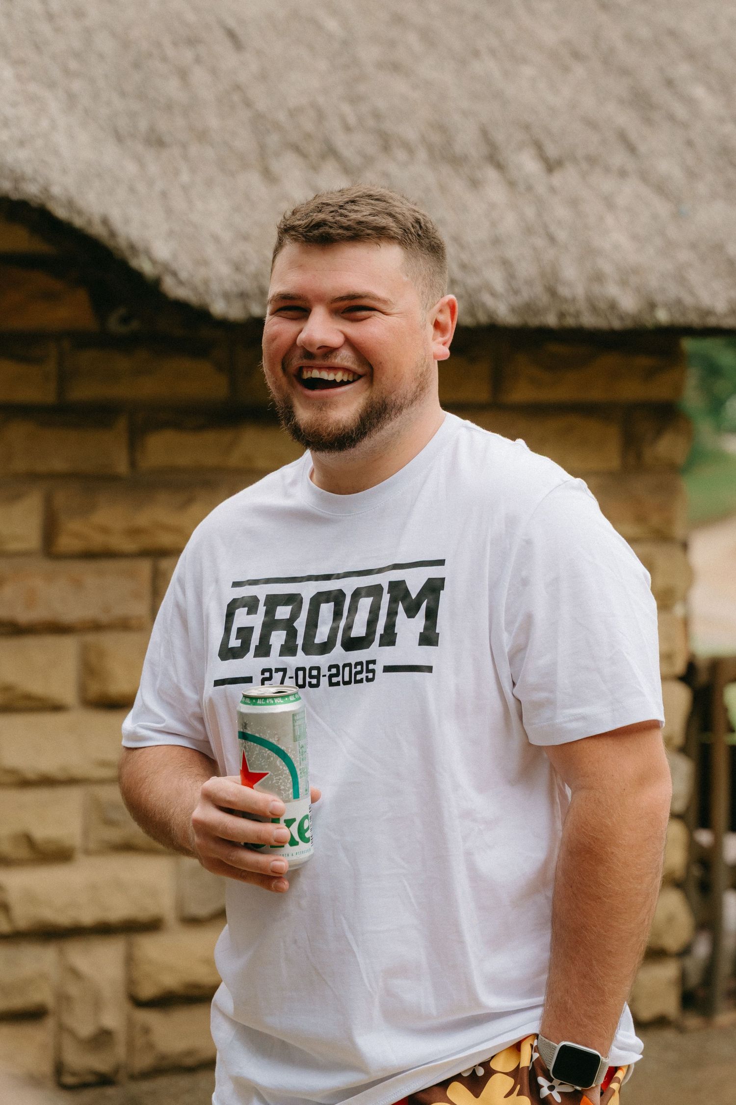 Wedding party member in white GROOM t-shirt holds a drink while laughing outdoors.