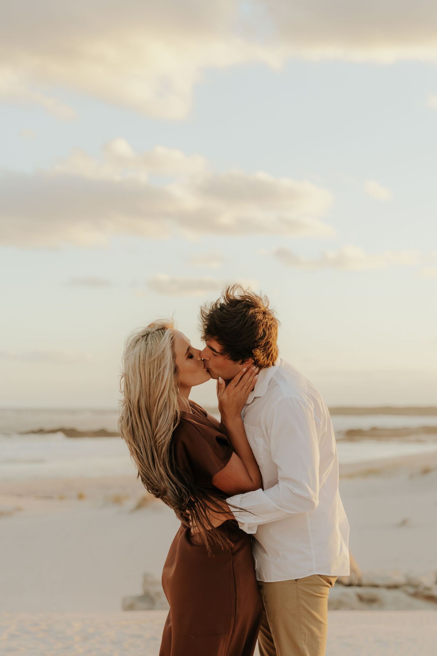 A couple shares a romantic kiss on a sandy beach at golden hour, the woman in a brown dress and man in white shirt.