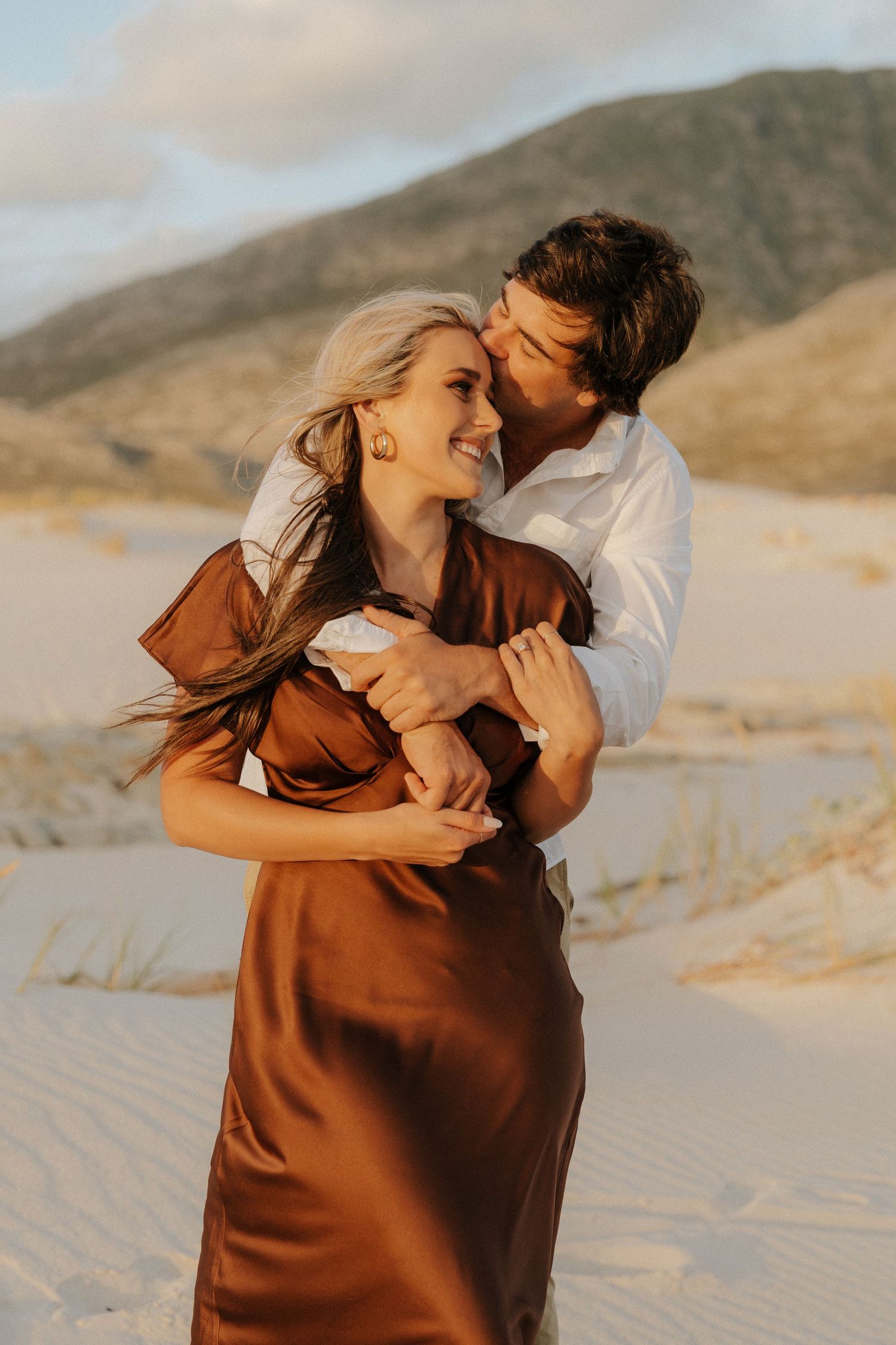A couple embraces on sandy dunes, woman in brown satin dress laughing as man in white shirt holds her close.