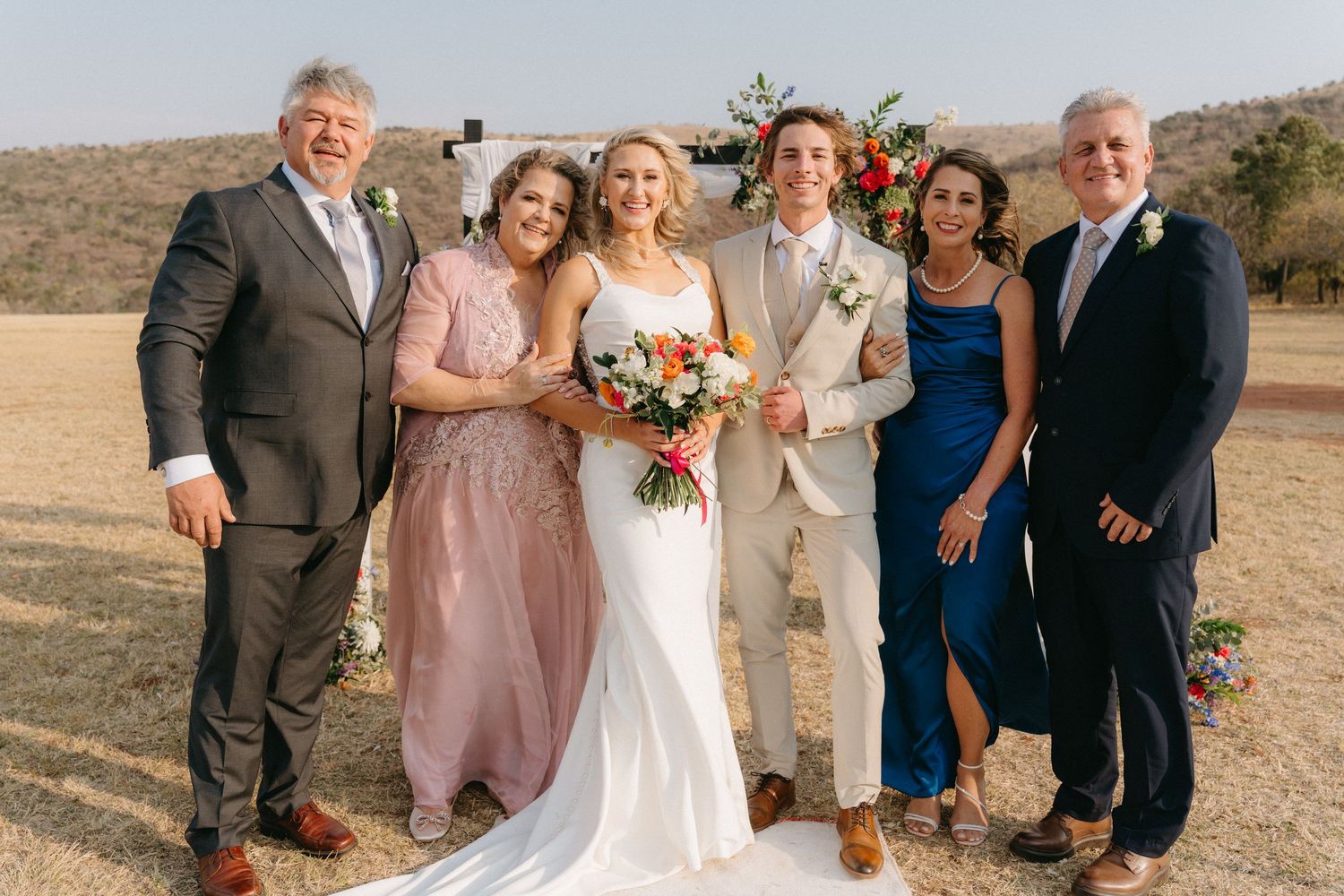 Wedding party poses for outdoor group photos in mix of formal attire against natural landscape.