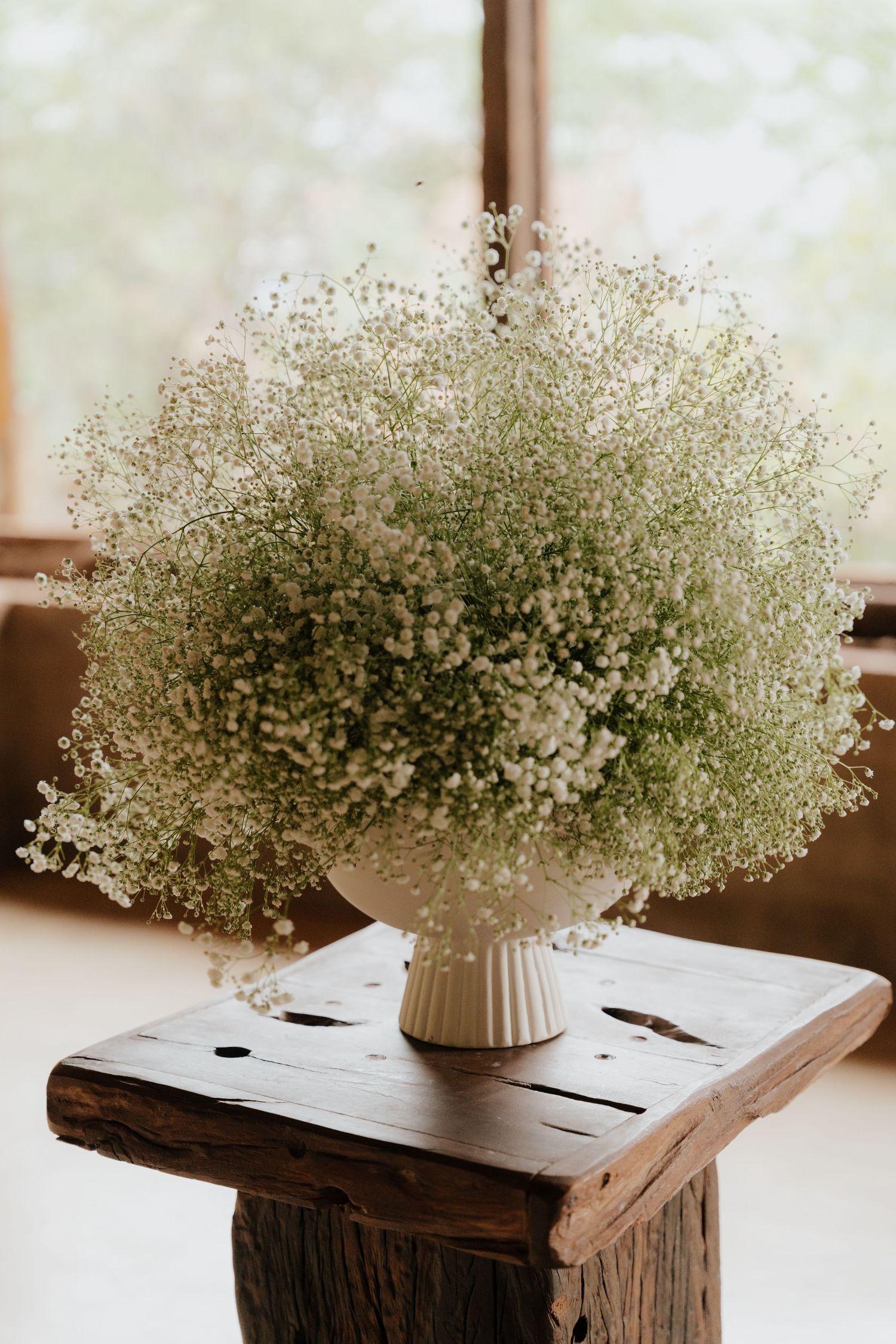 White baby's breath flowers arranged in a ribbed vase sit atop a rustic wooden surface near a window.