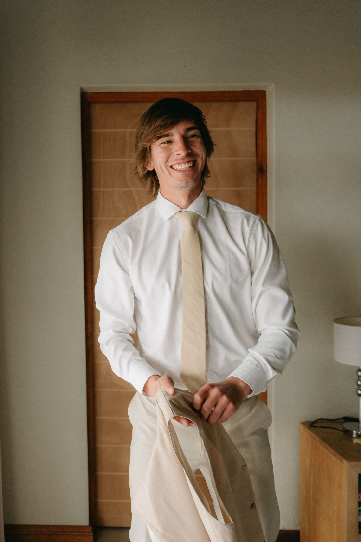 Person in formal attire adjusting their outfit while getting ready in a minimalist room with wooden doors.