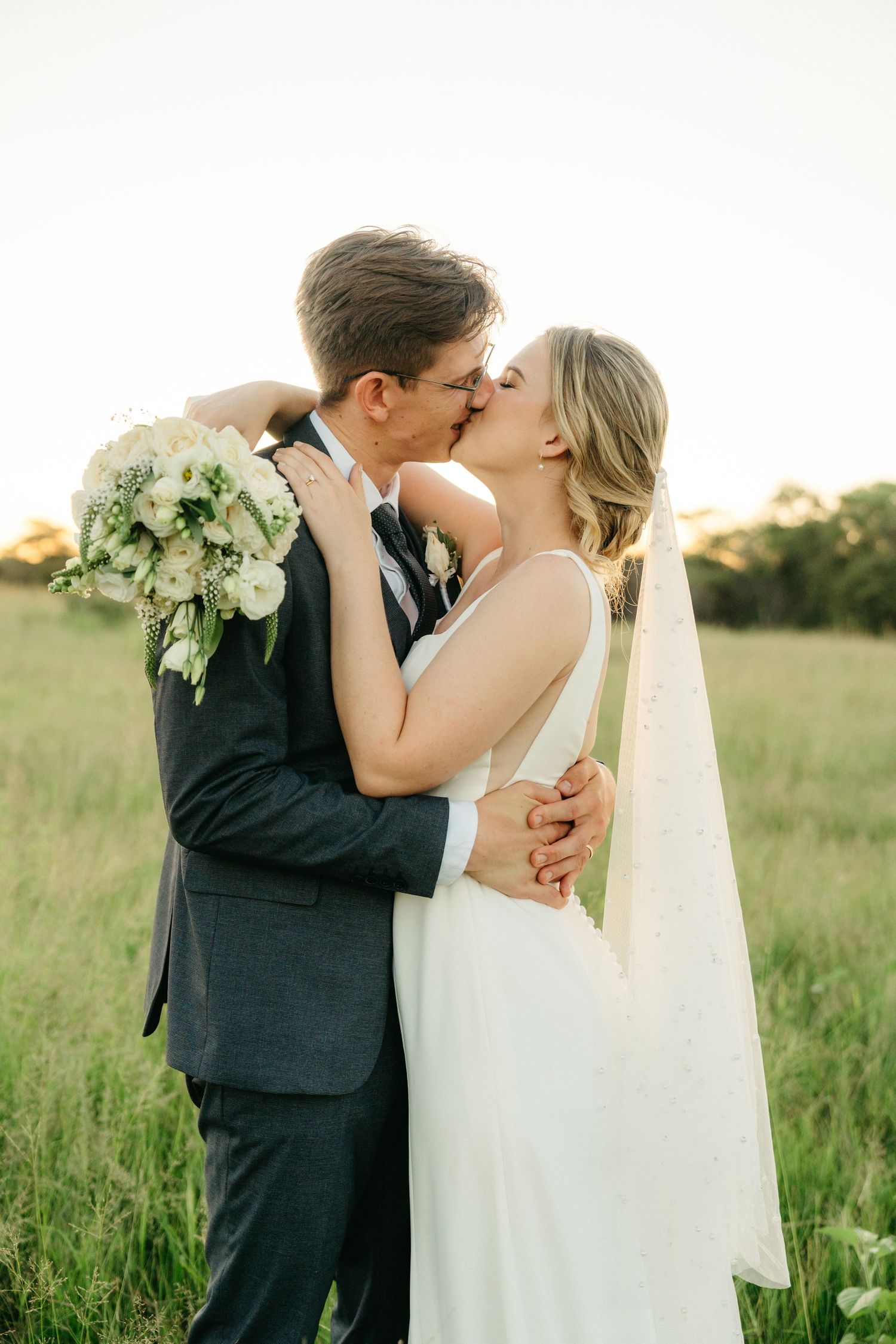 A romantic wedding portrait of a couple sharing a kiss in a sunlit meadow, with the bride holding a white floral bouquet.
