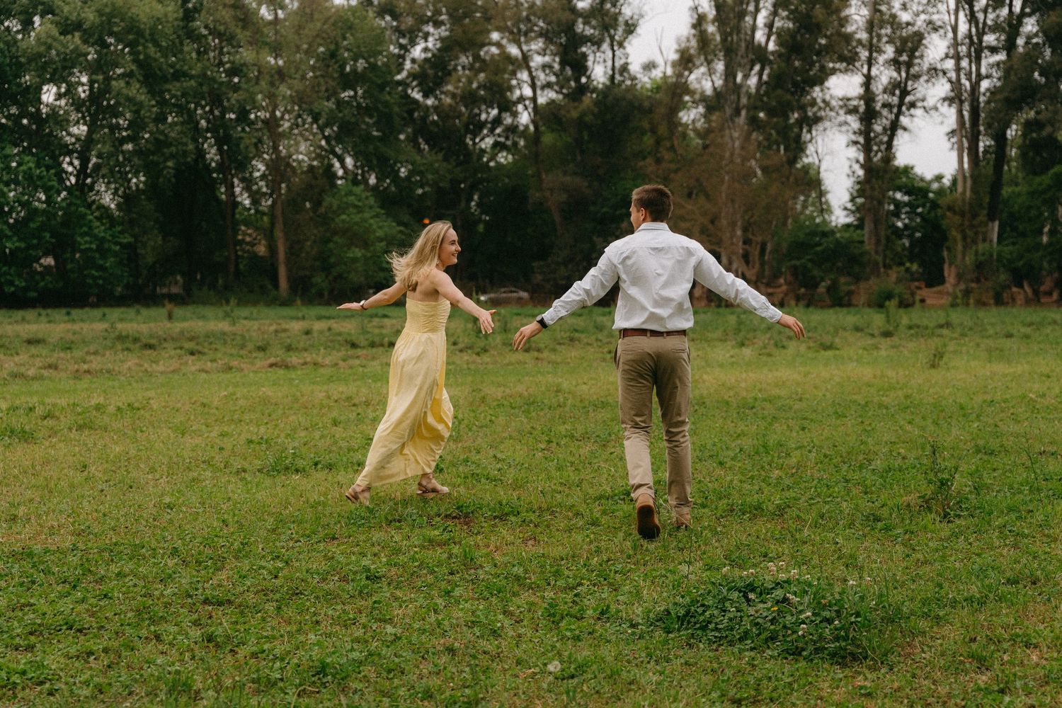Couple running hand in hand through green meadow with trees in background during engagement shoot at Irene Dairy Farm